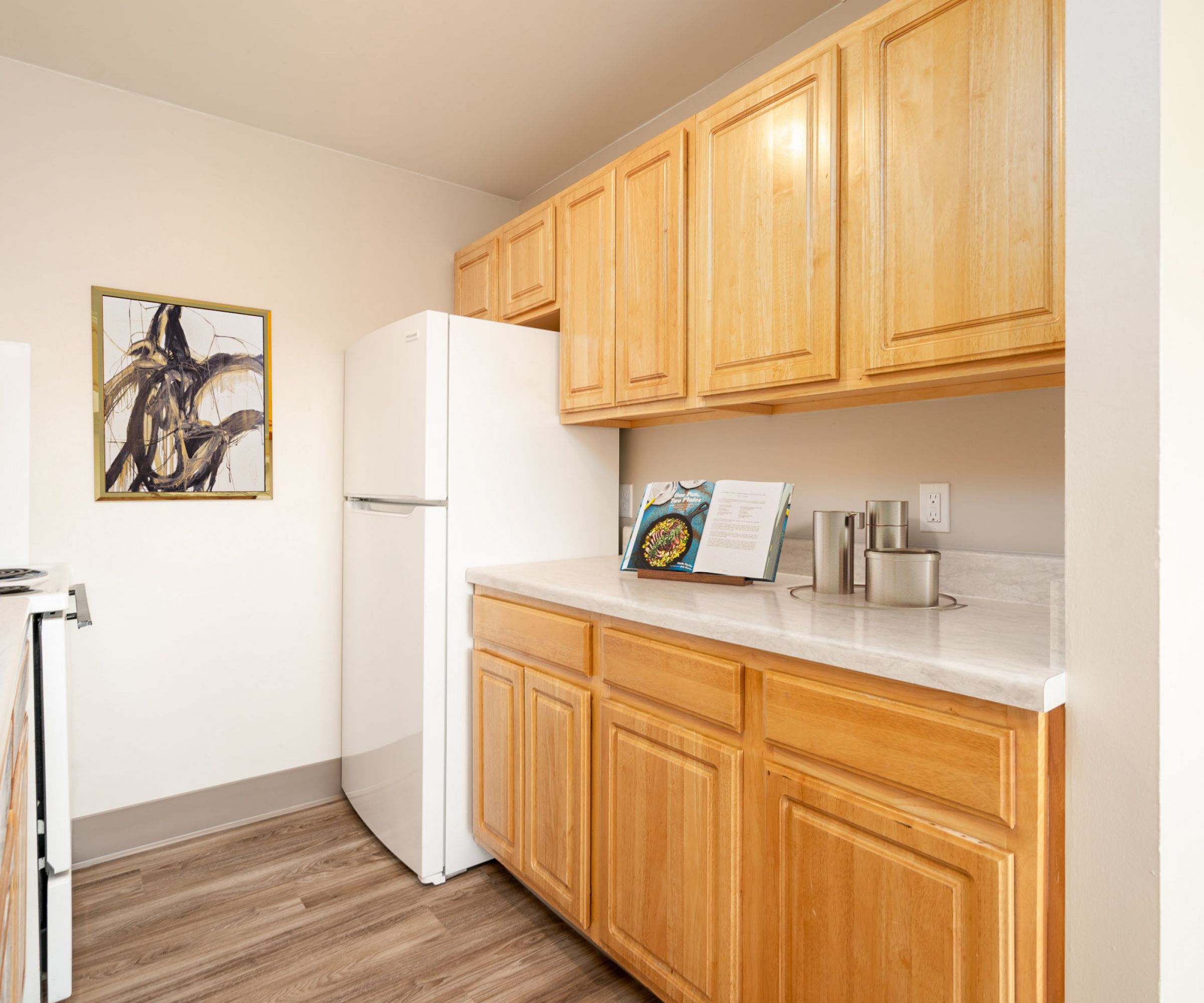 Aeon Affordable Homes and Community Affordable apartment homes feature a compact kitchen with wooden cabinets, a white fridge, a cookbook on the counter, and wall art on light wood floors.
