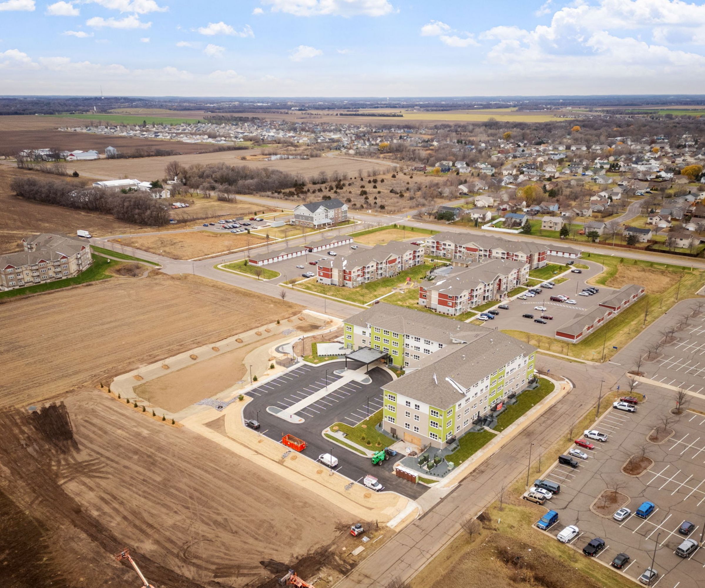 Aeon Affordable Homes and Community Aerial view of an affordable apartment homes complex surrounded by farmland and a parking lot under a partly cloudy sky.