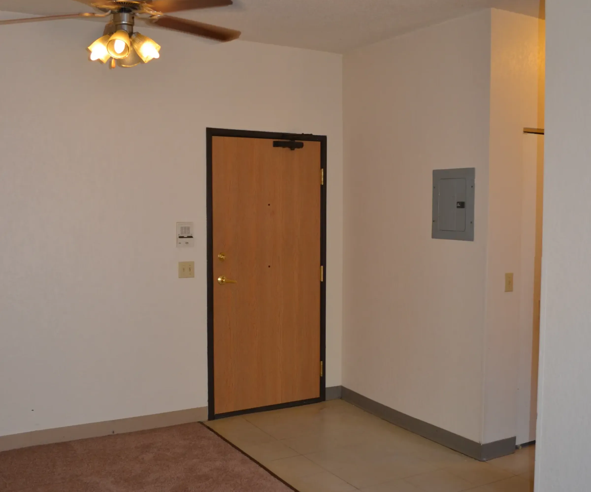 Aeon Affordable Homes and Community Affordable apartment homes featuring a room with a ceiling fan, beige carpet, and a wooden door, plus tile near the entryway and utility panel.