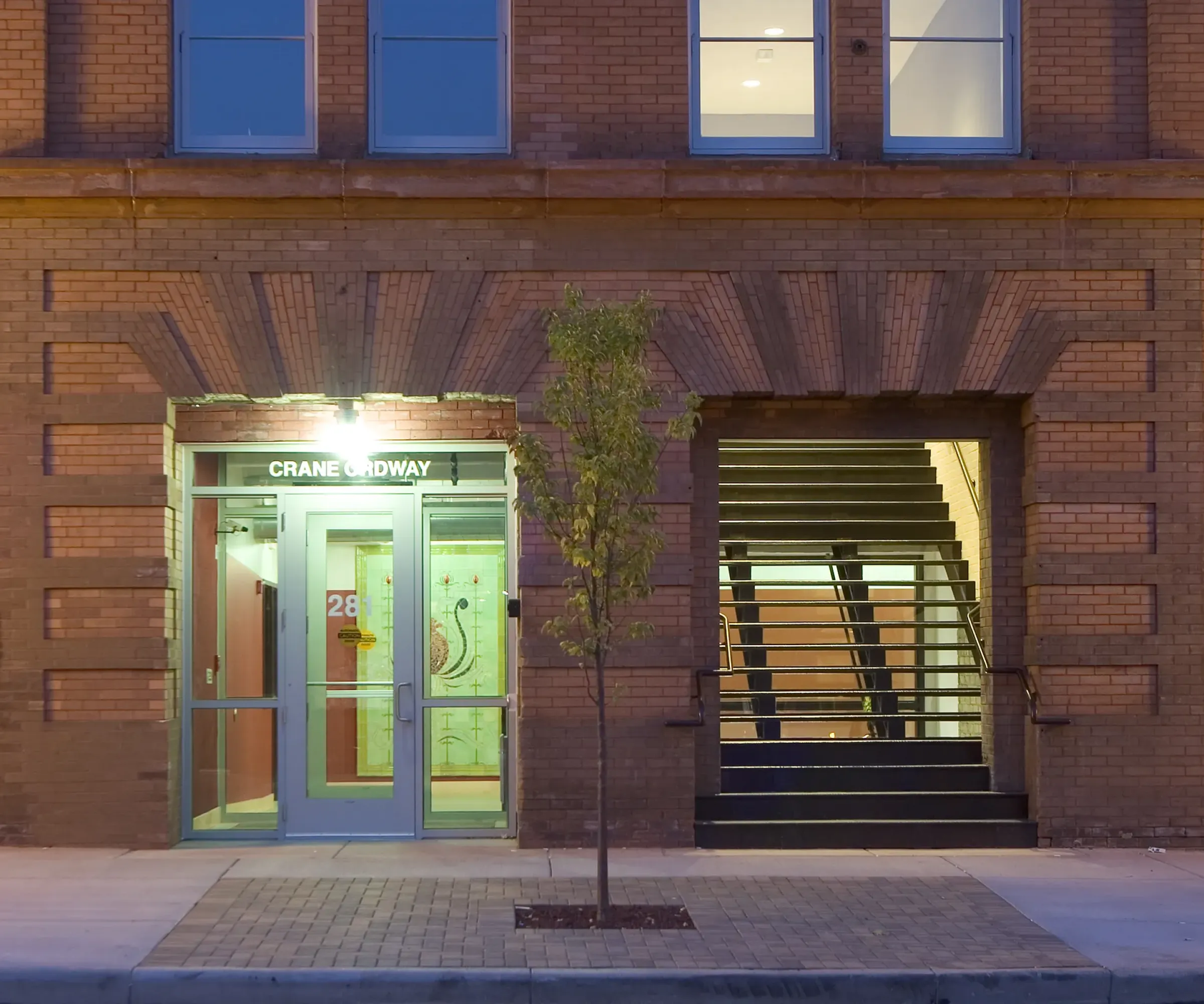 Aeon Affordable Homes and Community Affordable apartment homes with a brick facade, featuring a glass door entrance next to an open stairway, and a small tree on the sidewalk.