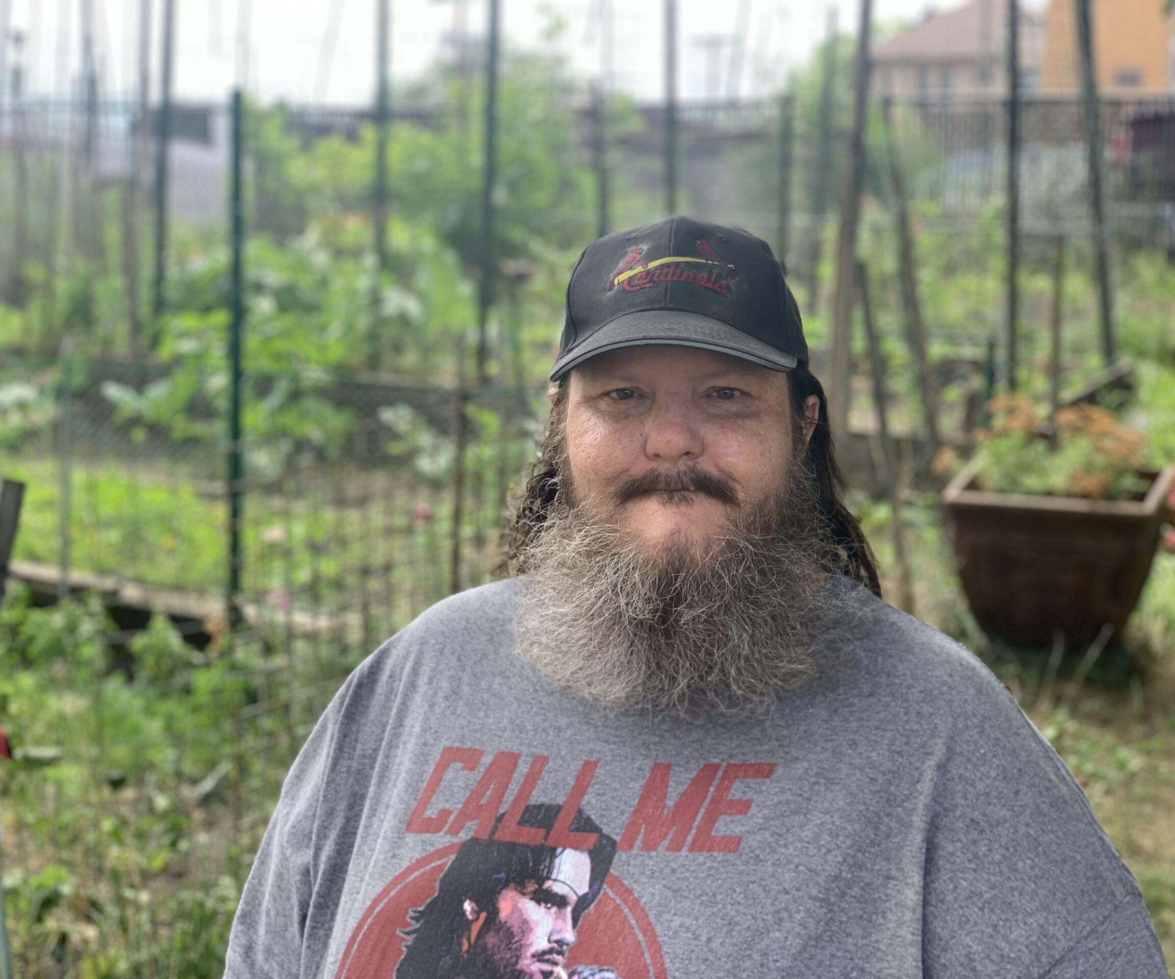 Aeon Affordable Homes Bearded man in a cap and graphic t-shirt stands in a lush garden, the perfect setting for Randy's Story among plants and trellises.