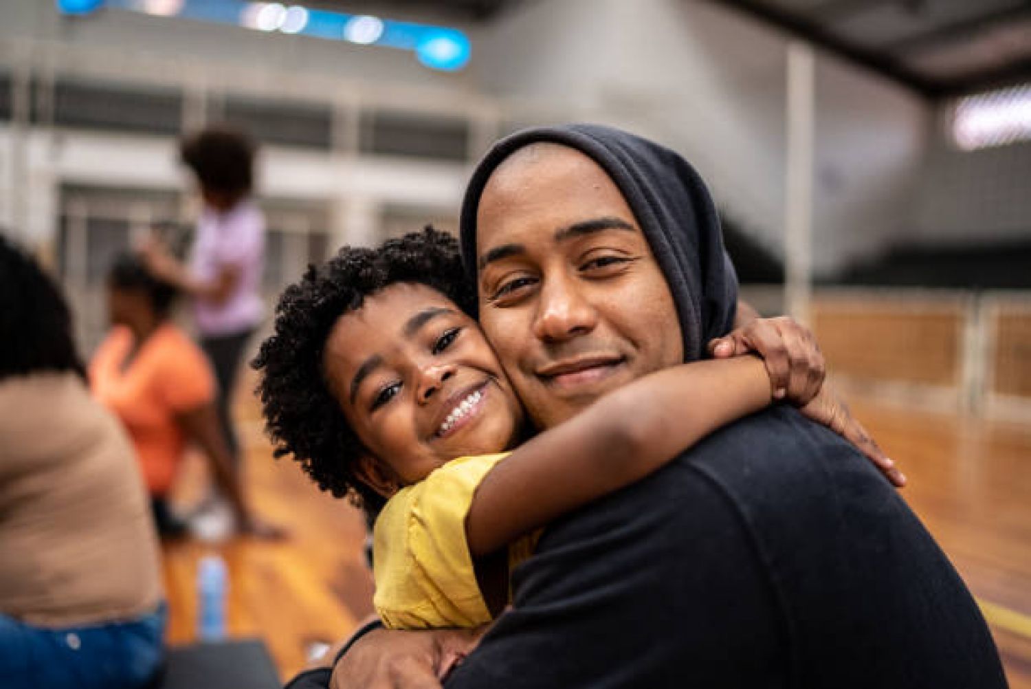Aeon Affordable Homes Smiling child hugging an adult in a hoodie at an indoor gym or sports facility.
