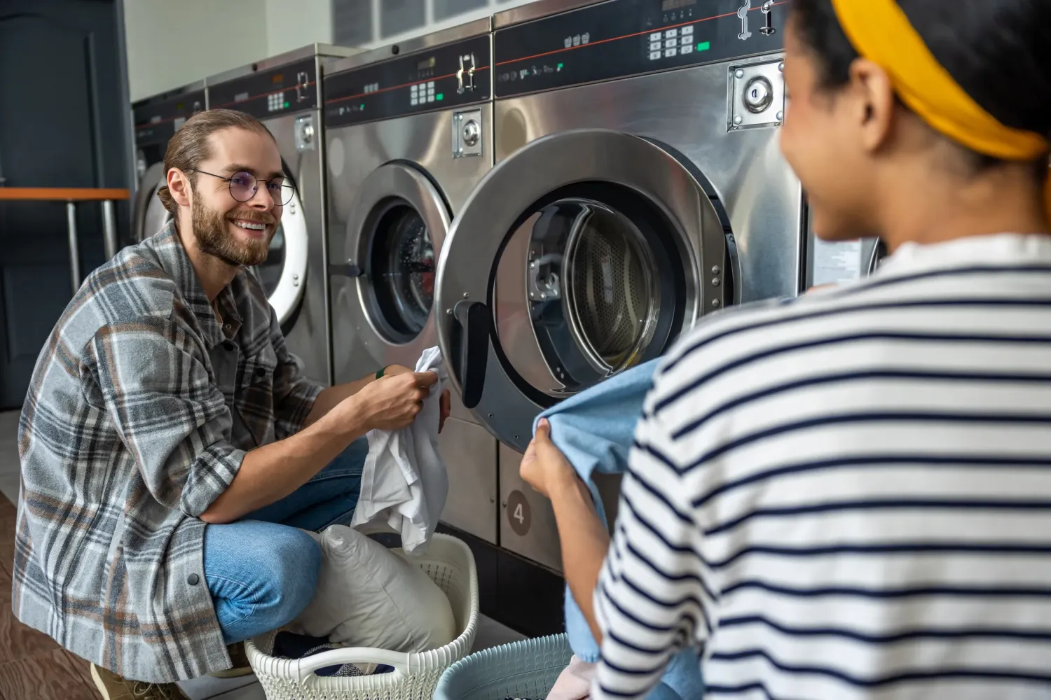 Aeon Affordable Homes with two people doing laundry at an apartment community shared laundry, smiling and folding clothes in front of industrial washing machines.