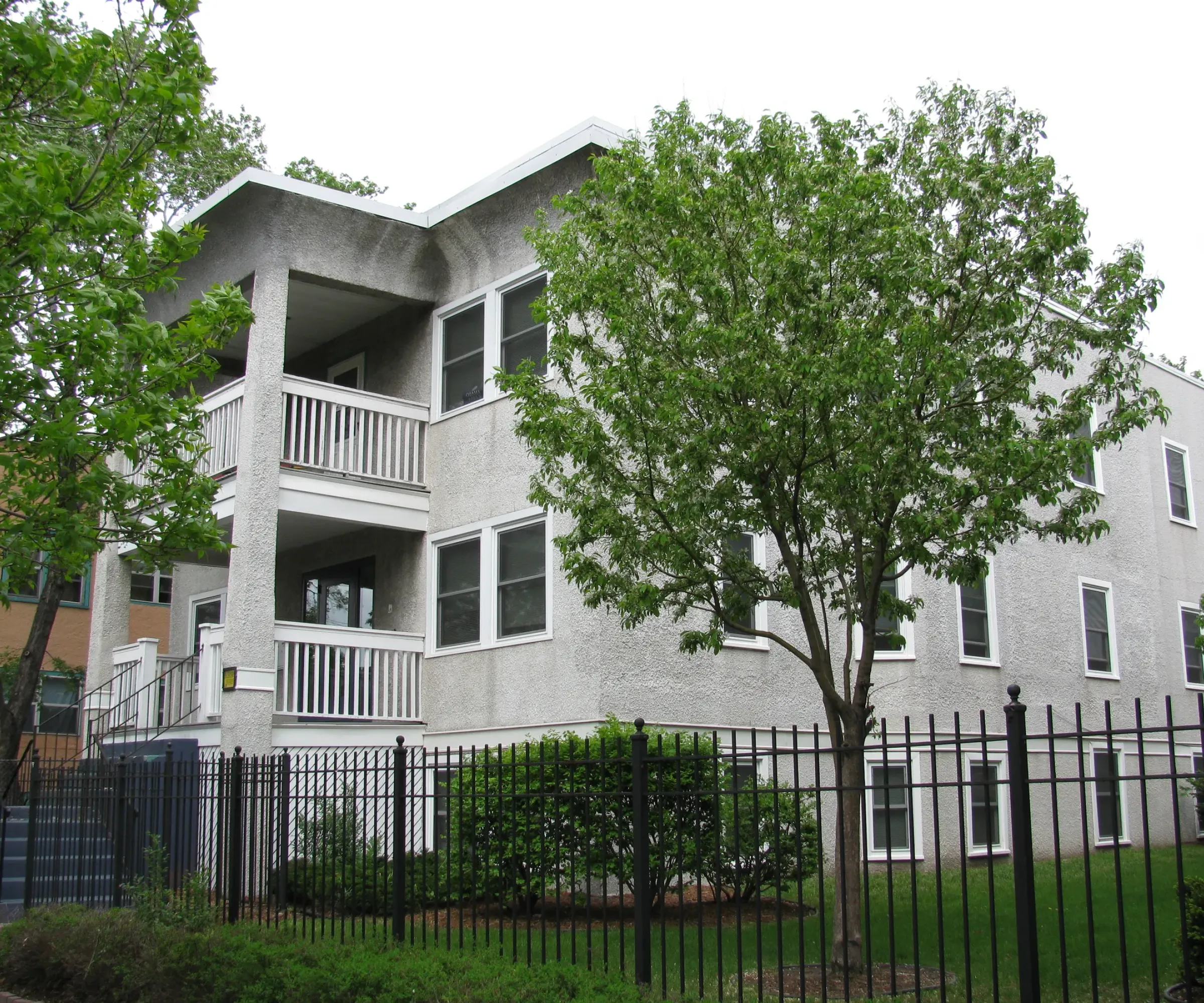 Aeon Affordable Homes and Community Affordable apartment homes in a two-story gray building, complete with balconies, surrounded by trees and a black fence.
