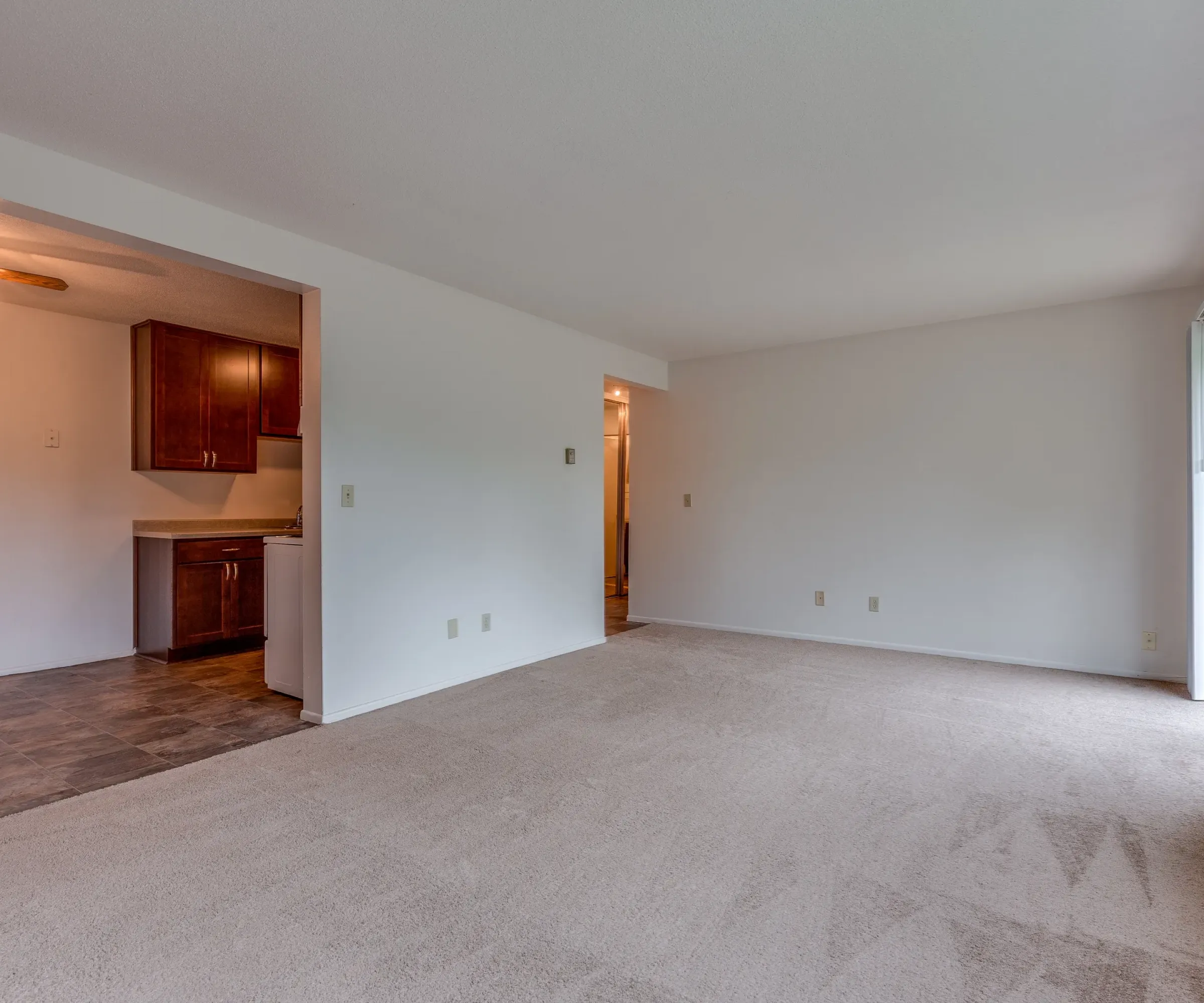 Aeon Affordable Homes and Community Empty living room with beige carpet in an affordable apartment home, adjacent to a kitchen with dark cabinets and a ceiling fan; sliding glass door on right.