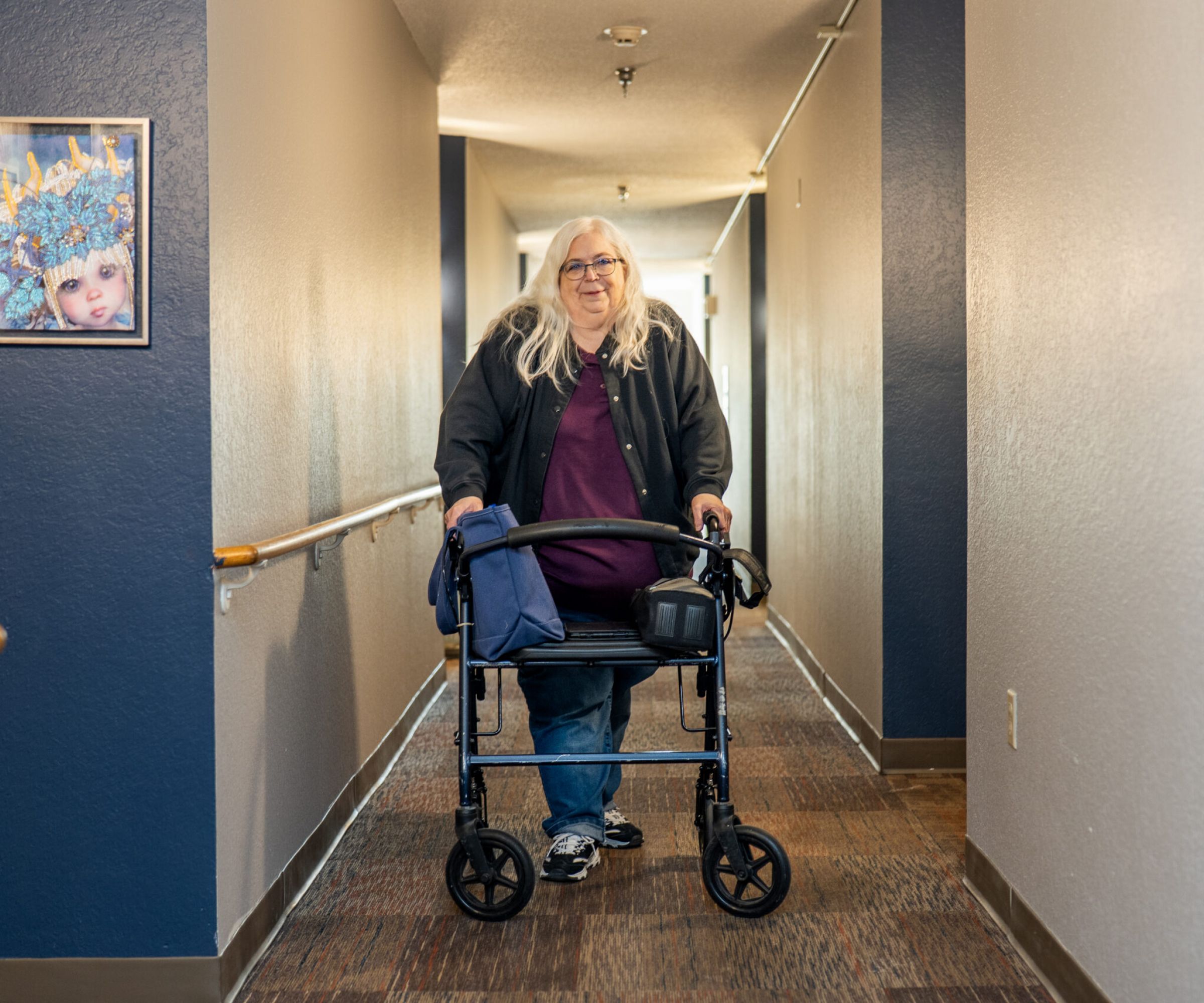 Aeon Affordable Homes An older woman with long white hair uses a walker in a hallway, smiling at the camera—this is part of Dianne's Story.