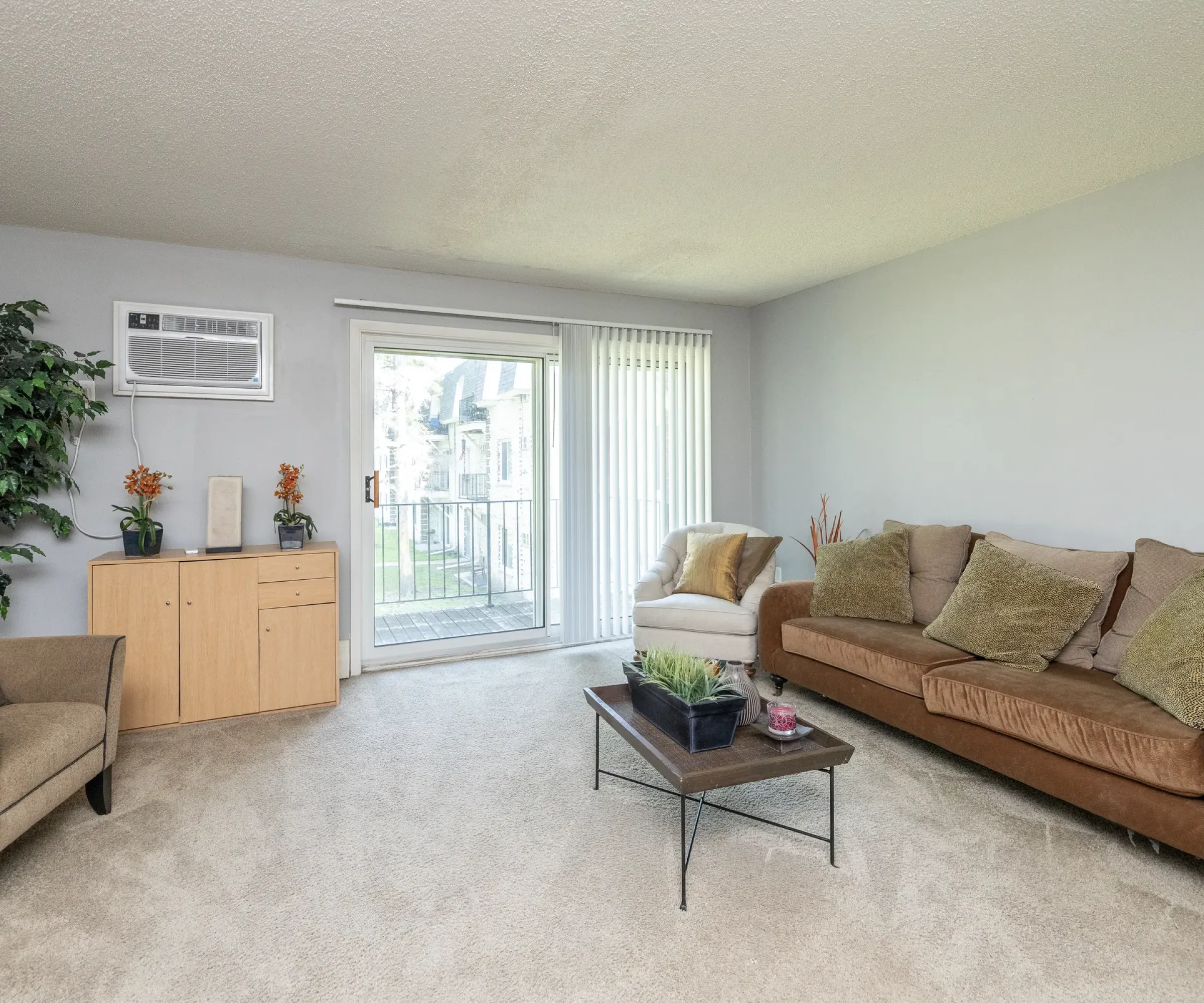 Aeon Affordable Homes Living room with a brown sofa, armchair, coffee table, and plants; enjoy a Blooming Meadows South balcony view through sliding glass doors.