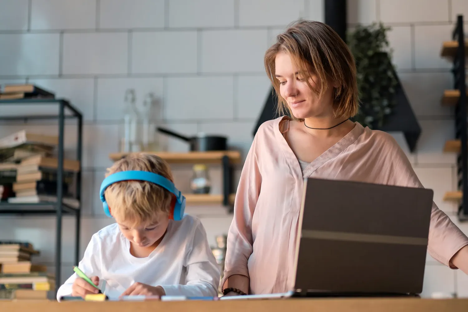 Aeon Affordable Homes with a woman standing behind a laptop smiling at a boy in blue headphones doing homework in a kitchen.