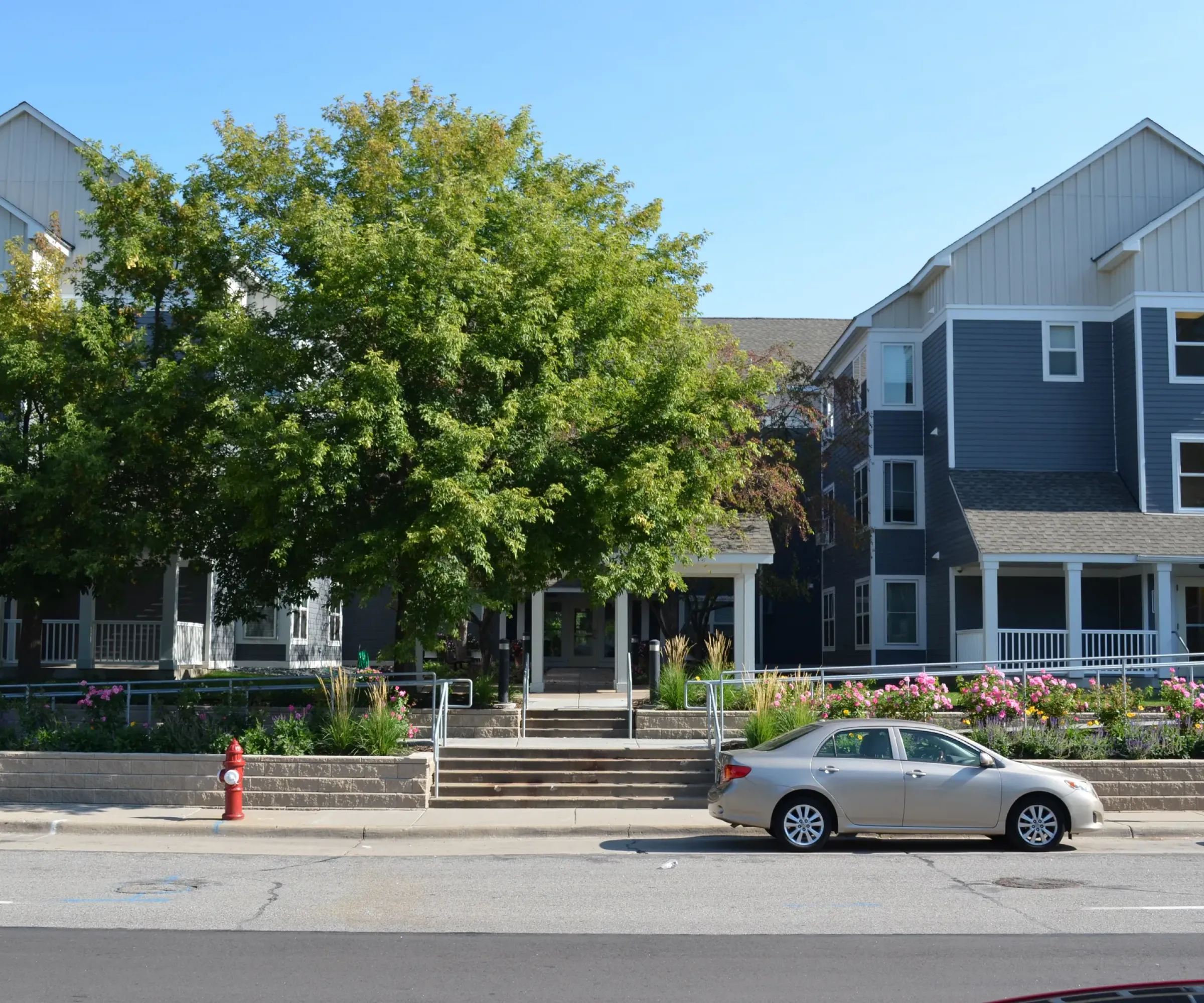 Aeon Affordable Homes and Community Affordable apartment homes feature gray and white siding, lush trees, and a parked silver car in front.