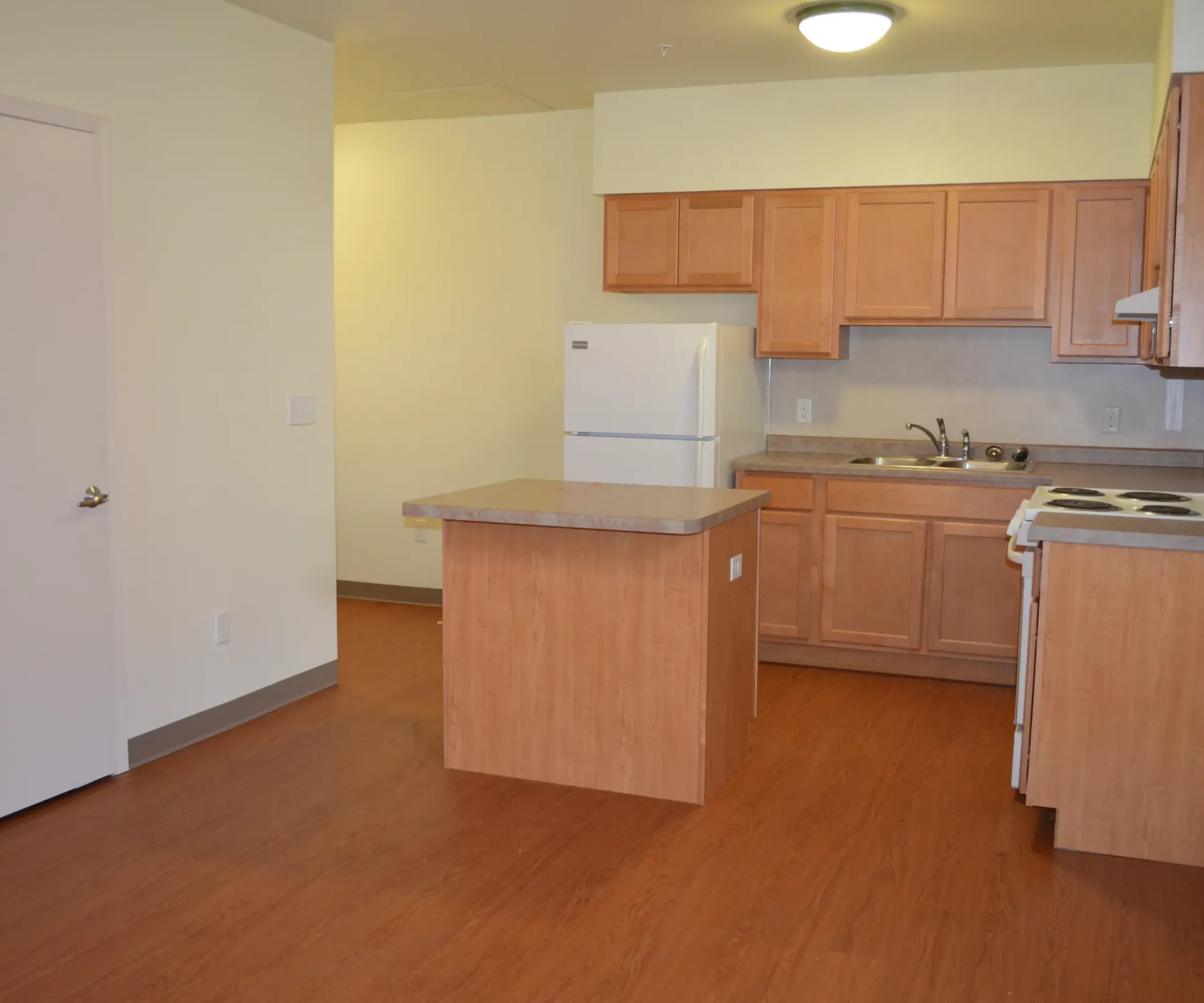 Aeon Affordable Homes and Community A cozy kitchen in affordable apartment homes with wooden cabinets, an island, a white refrigerator and stove on a brown floor. White walls and door.