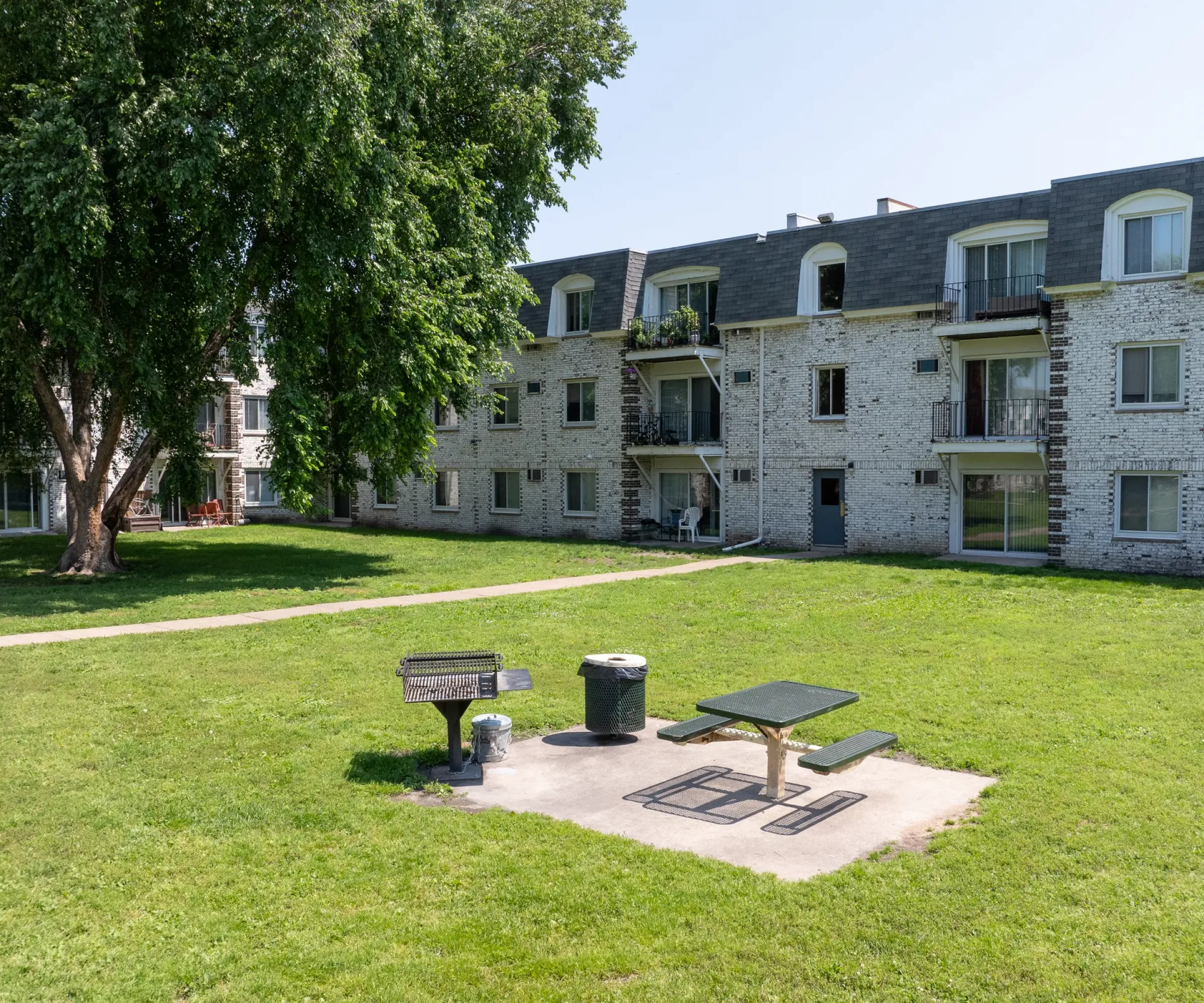 Aeon Affordable Homes Grass courtyard at Blooming Meadows South with a picnic table, barbecue grill, and trash can in front of a three-story brick apartment building.