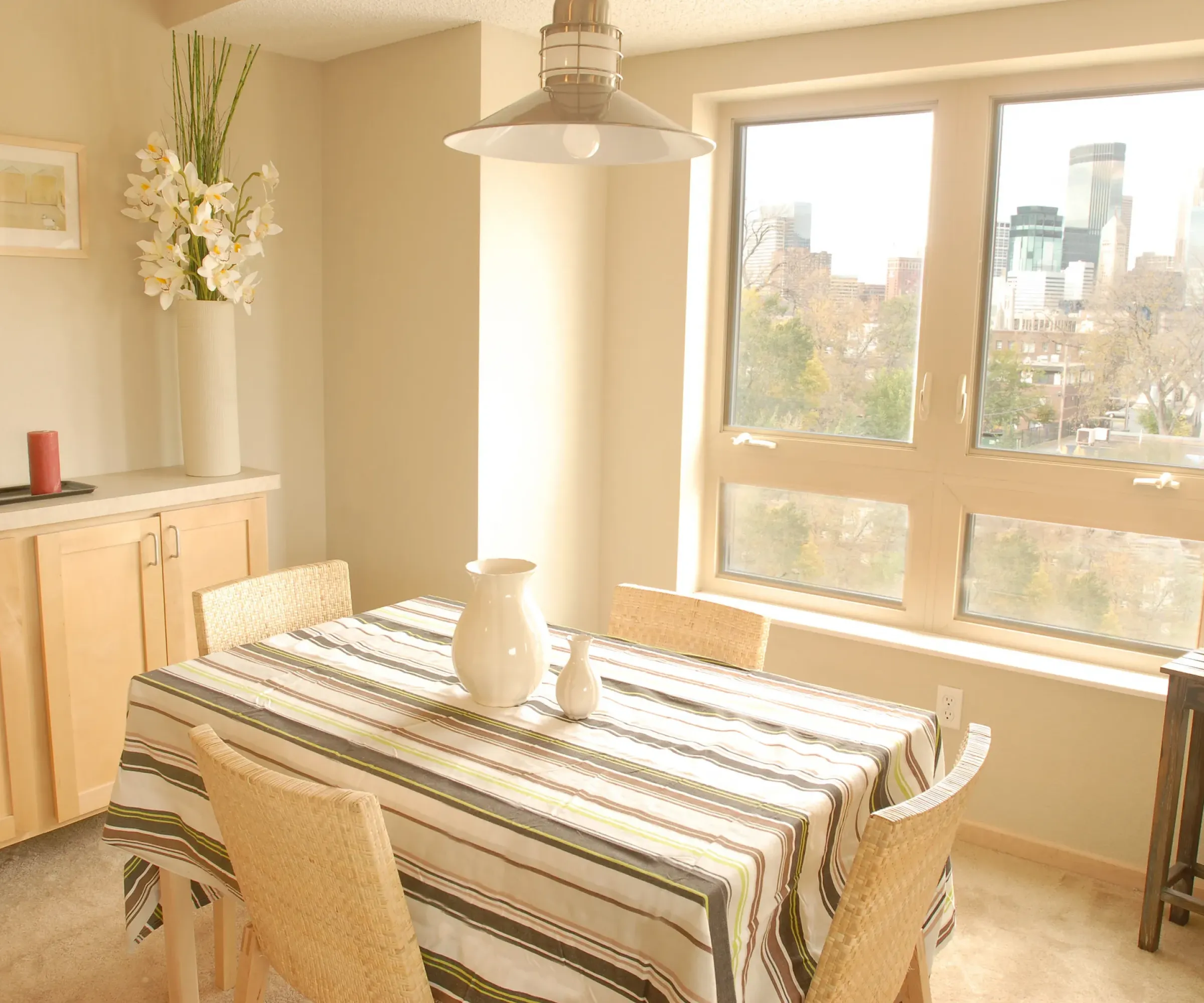 Aeon Affordable Homes and Community A dining room in affordable apartment homes, featuring a striped tablecloth, wicker chairs, cityscape view, and decorative plants.
