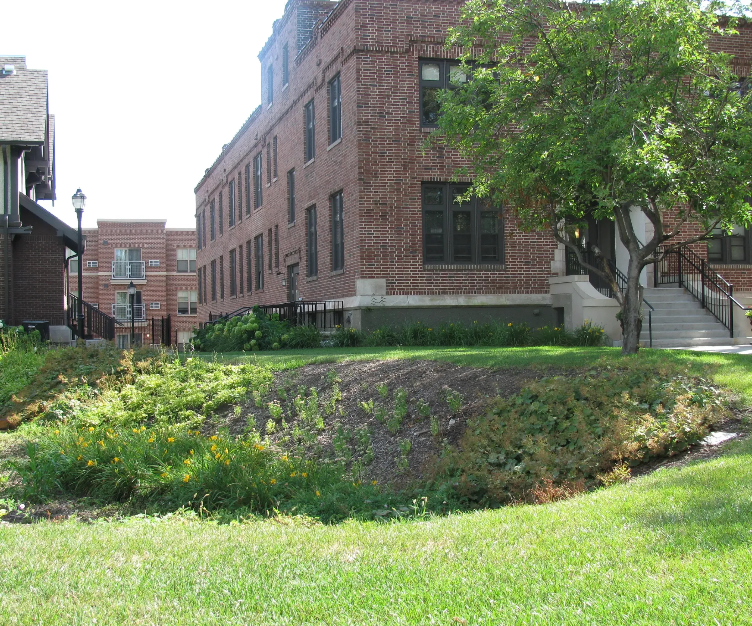 Aeon Affordable Homes and Community Affordable apartment homes in a brick building with a sloped green garden, featuring flowers, shrubs, and a tree near the entrance.