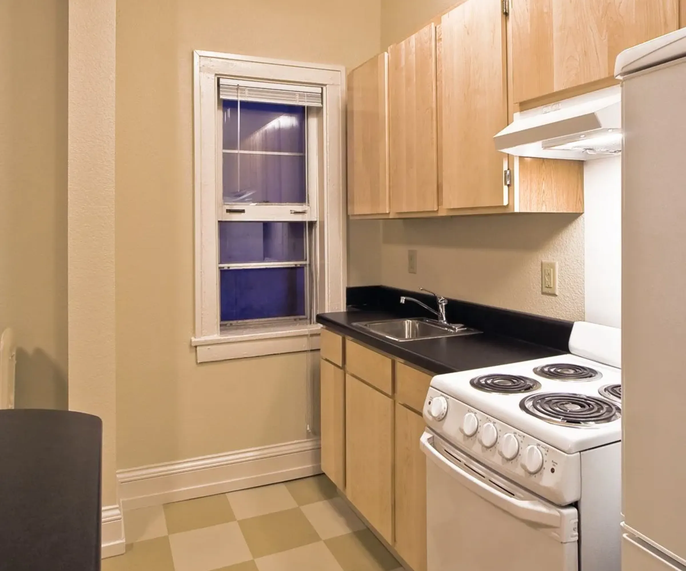 Aeon Affordable Homes Small kitchen with a white stove, fridge, wooden cabinets, black countertop, and checkered floor. Window above sink.