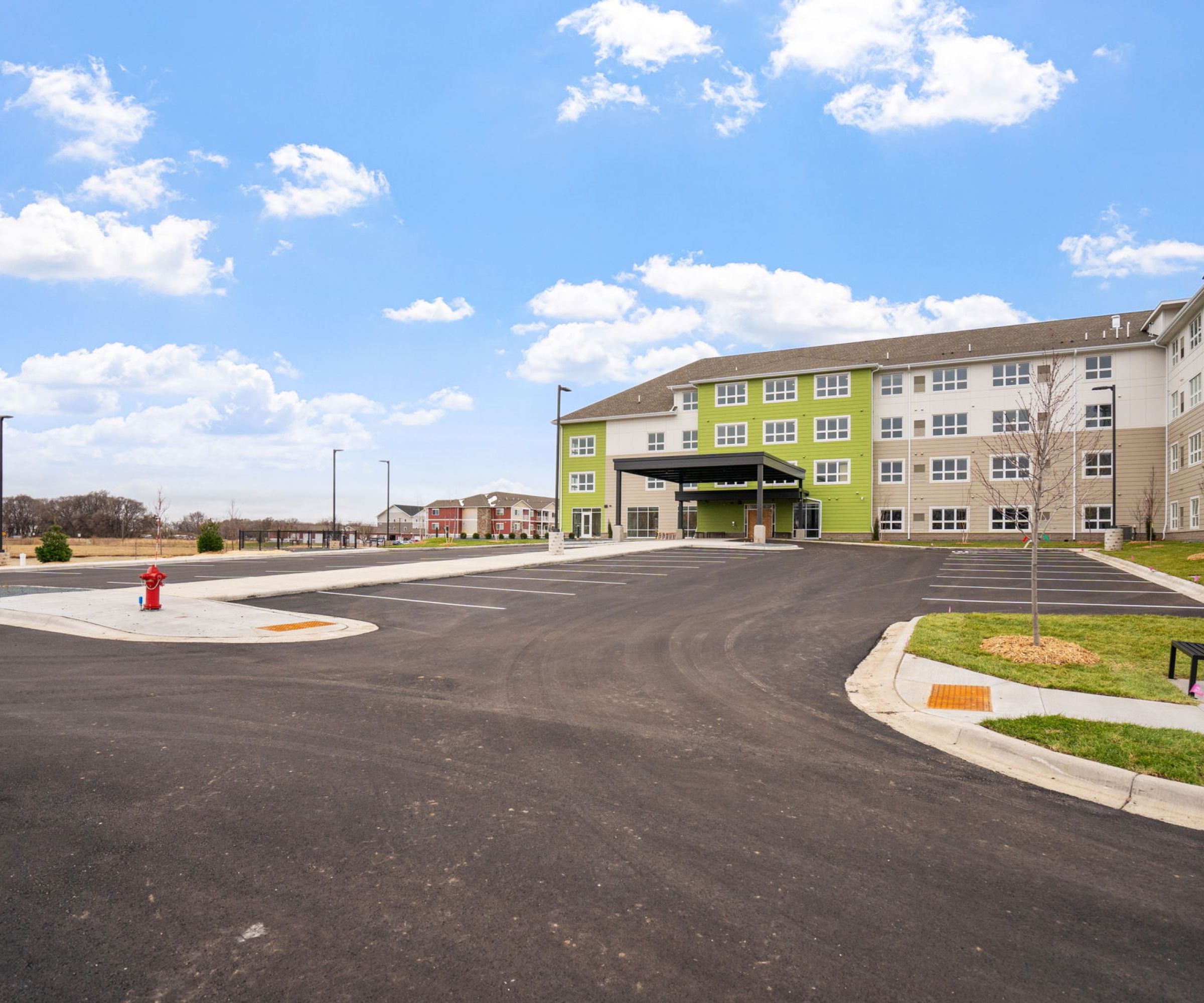 Aeon Affordable Homes and Community Modern multi-story building with green accents offering affordable apartment homes, large parking area, and blue sky with clouds.