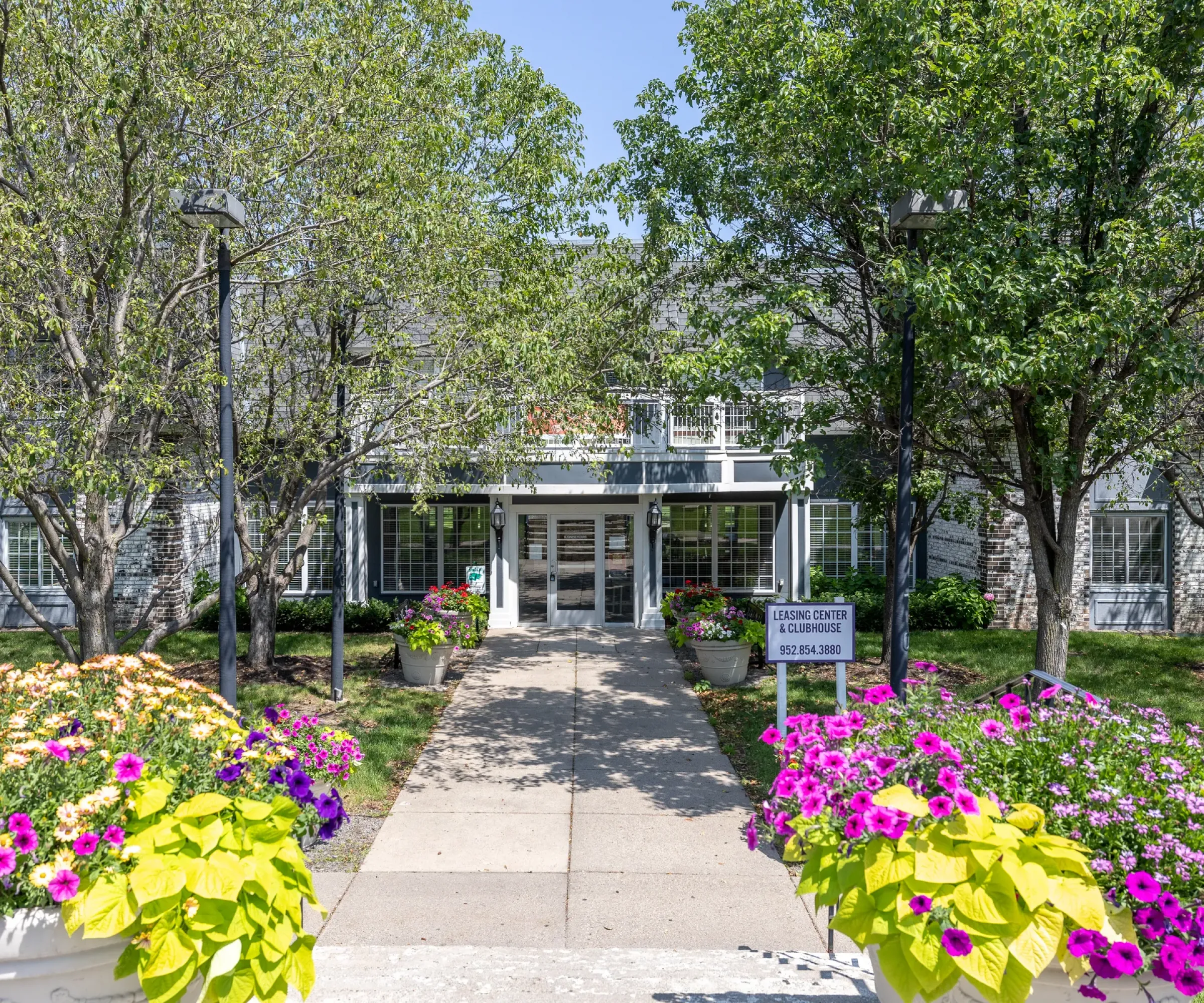 Aeon Affordable Homes A flower-lined path leads to the Lyon Center, nestled among trees under a clear sky, reminiscent of the blooming meadows of the South.