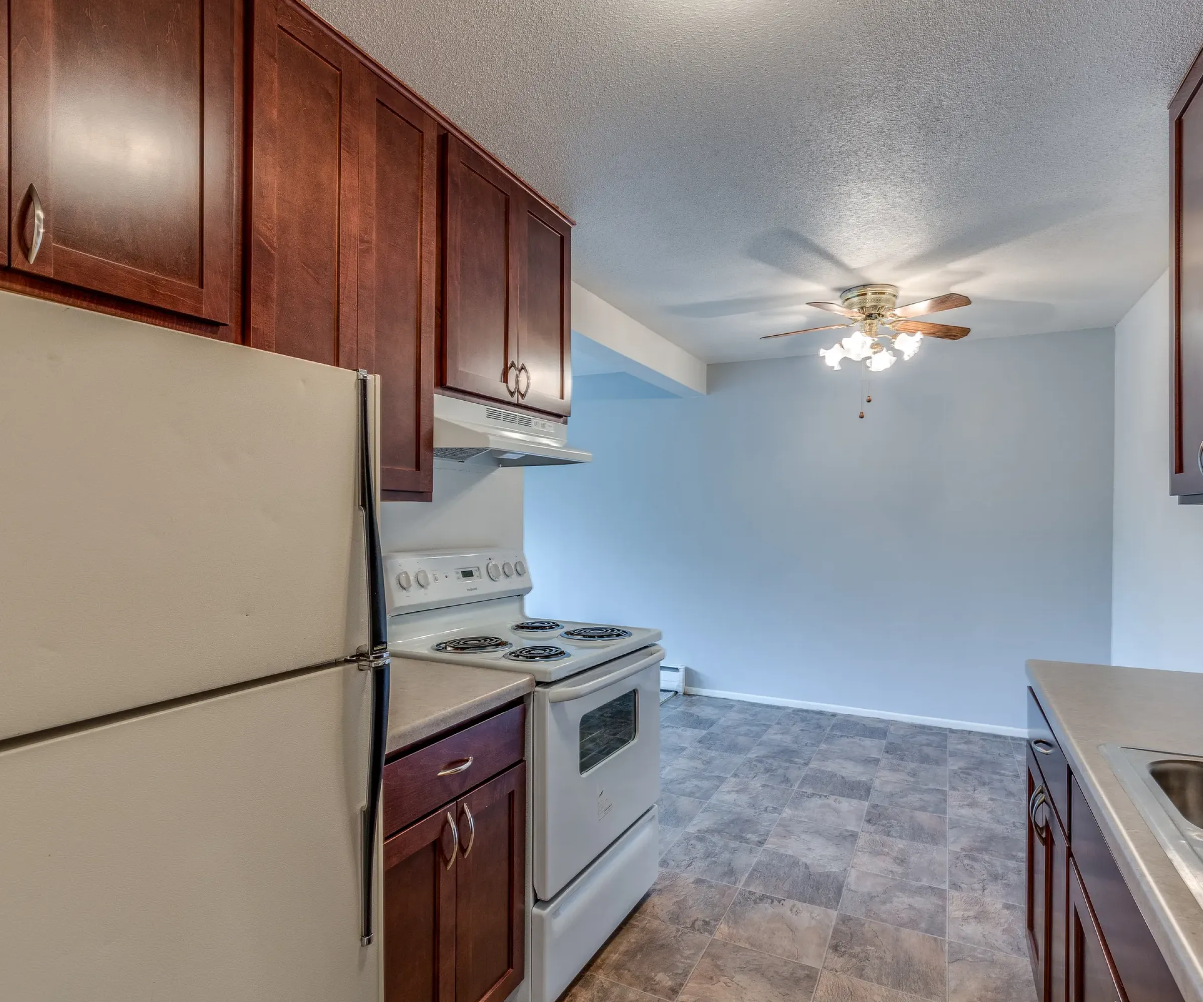 Aeon Affordable Homes and Community Affordable apartment homes with kitchen featuring dark wood cabinets, white appliances, and beige countertops; ceiling fan in dining area.