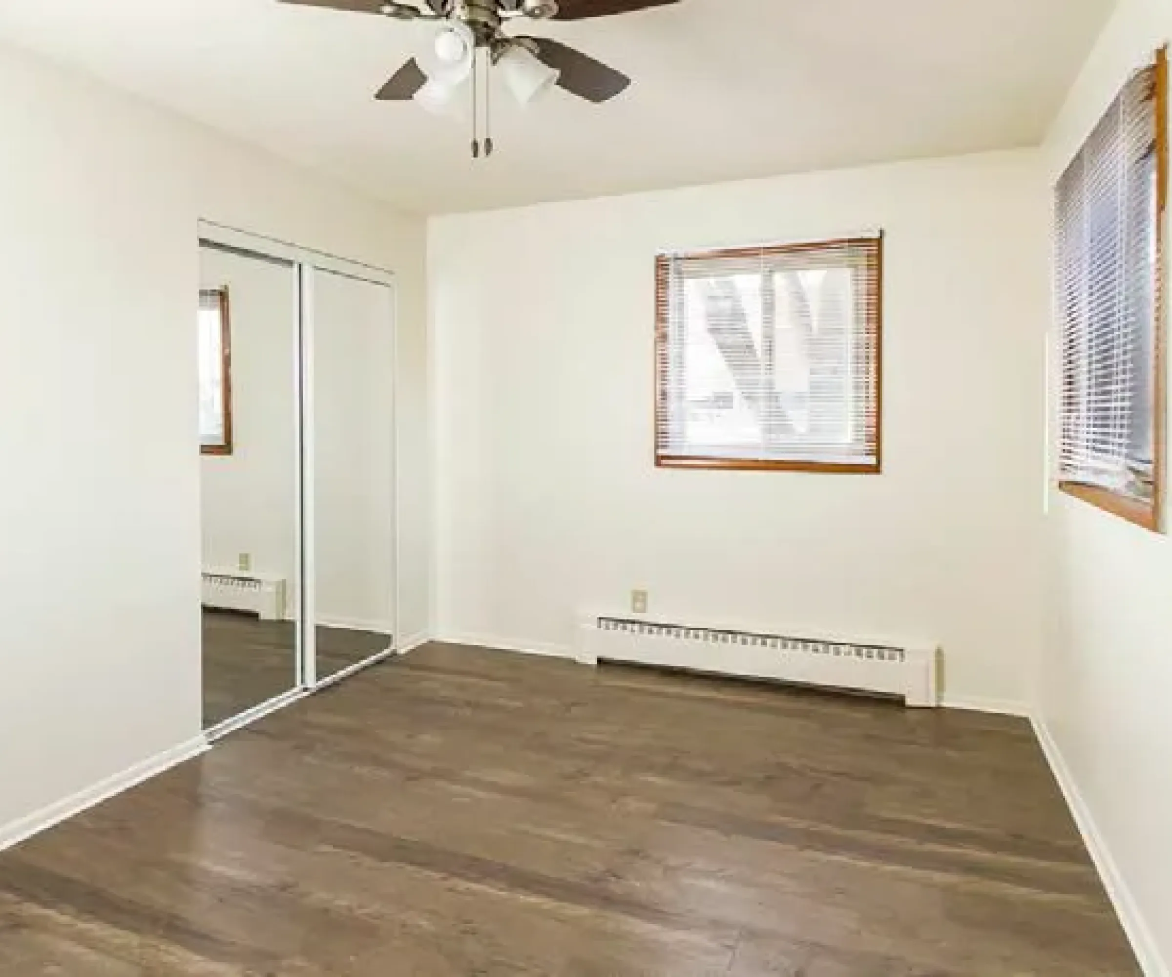 Aeon Affordable Homes Empty room with wooden floor, white walls, ceiling fan, and mirrored closet in Cobblestone Court apartments. Two windows with blinds on side walls.