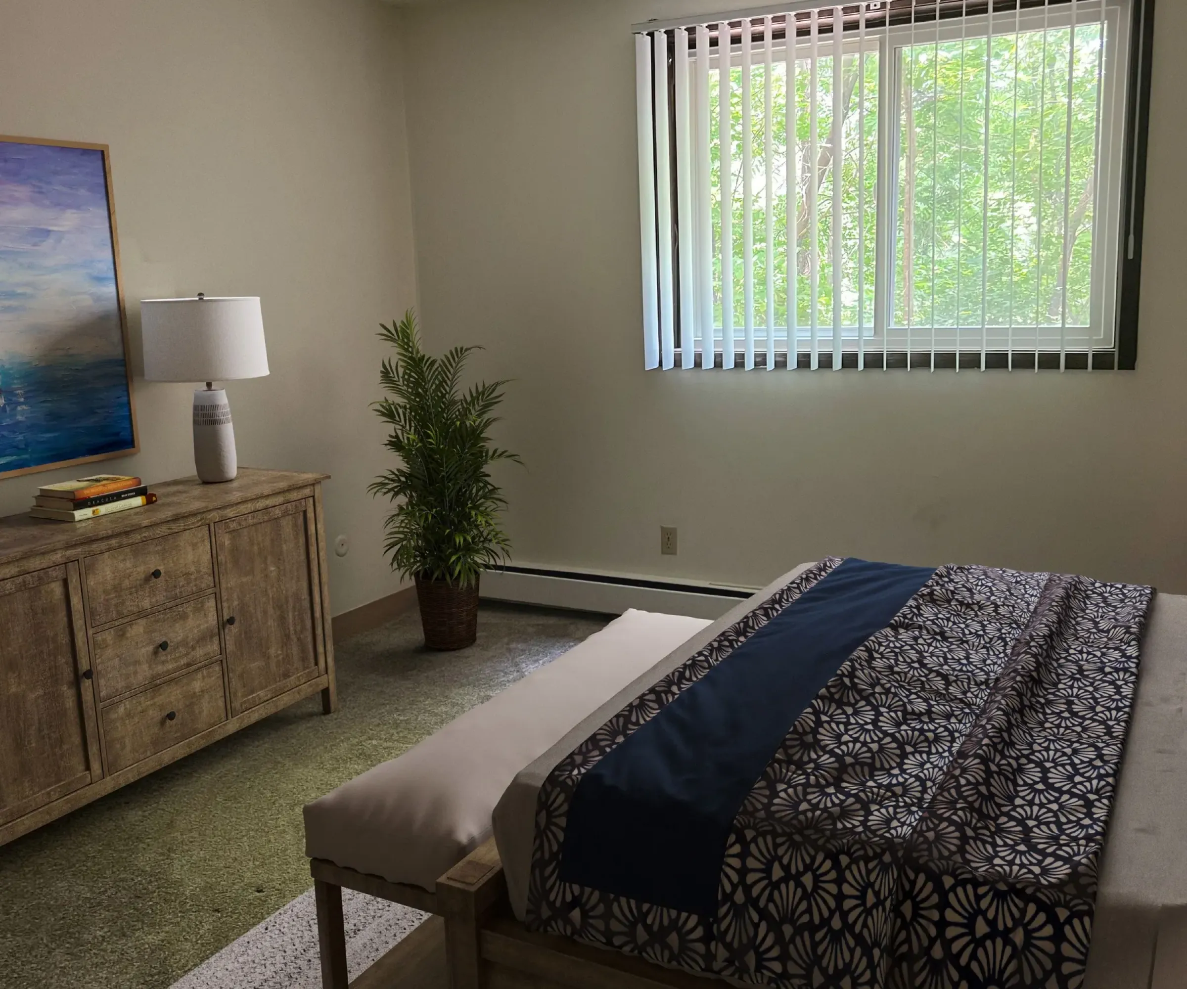 Aeon Affordable Homes and Community An affordable apartment bedroom featuring a bed, dresser, lamp, potted plant, and window with vertical blinds letting in natural light.