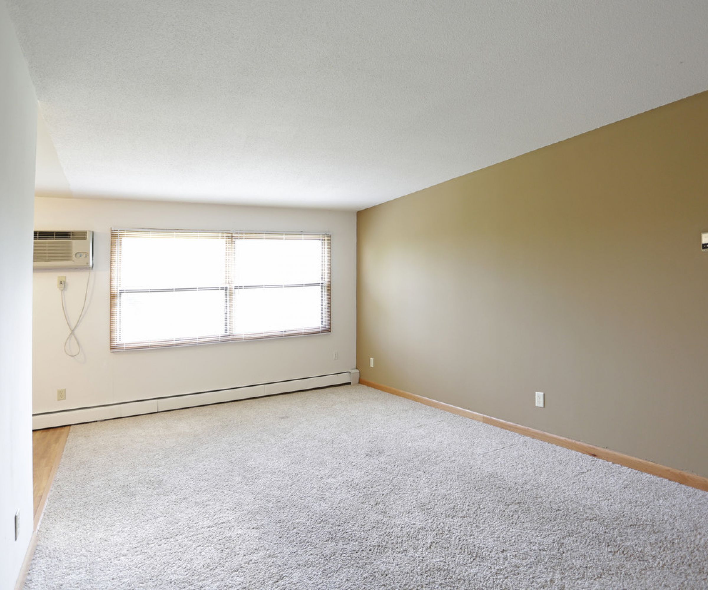 Aeon Affordable Homes and Community Affordable apartment homes featuring an empty room with beige carpet, tan walls, a large window, and a wall-mounted air conditioner.