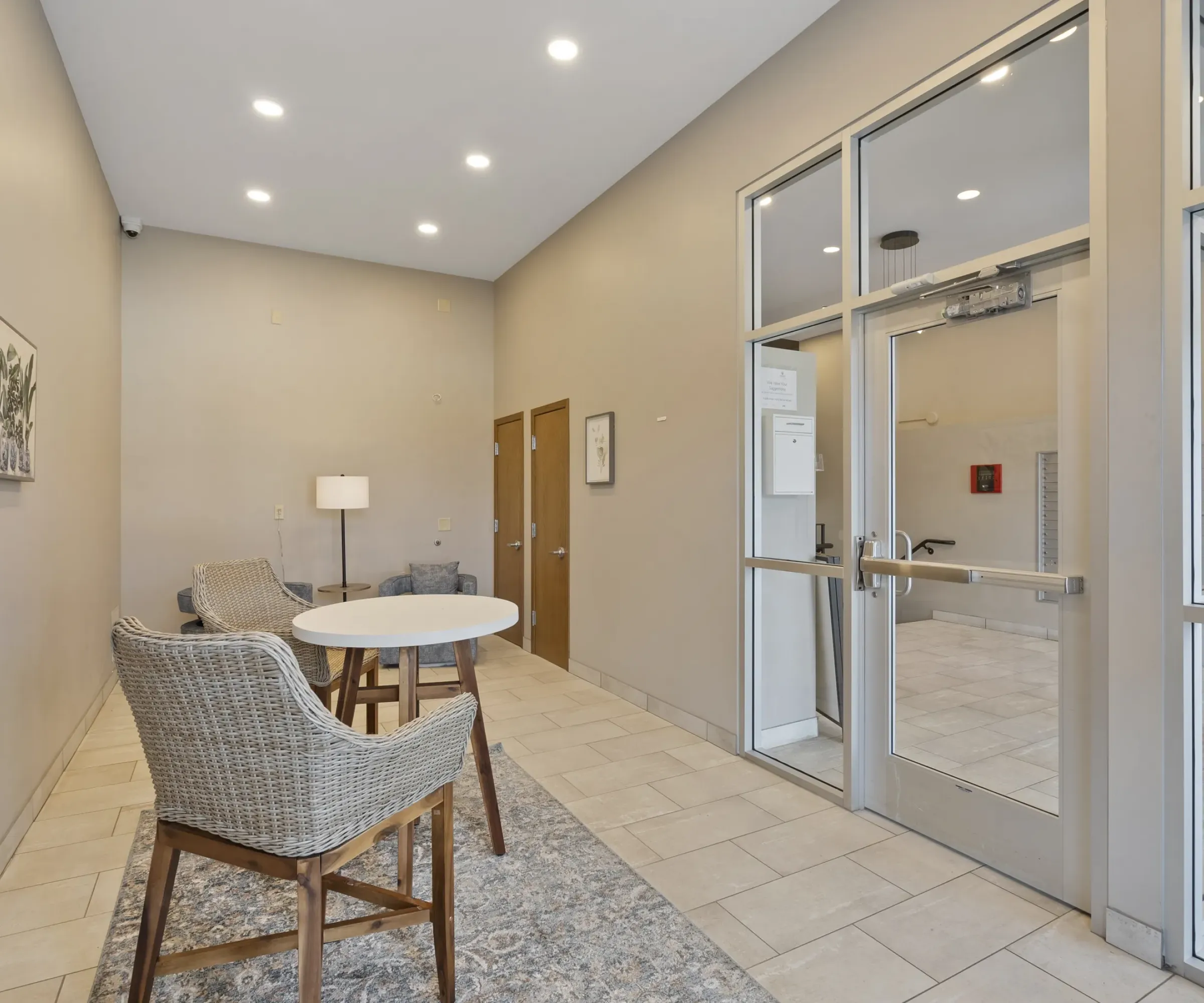 Aeon Affordable Homes Modern lobby with wicker chairs, a round table, and a lamp on a rug. Glass doors lead to an adjacent area.