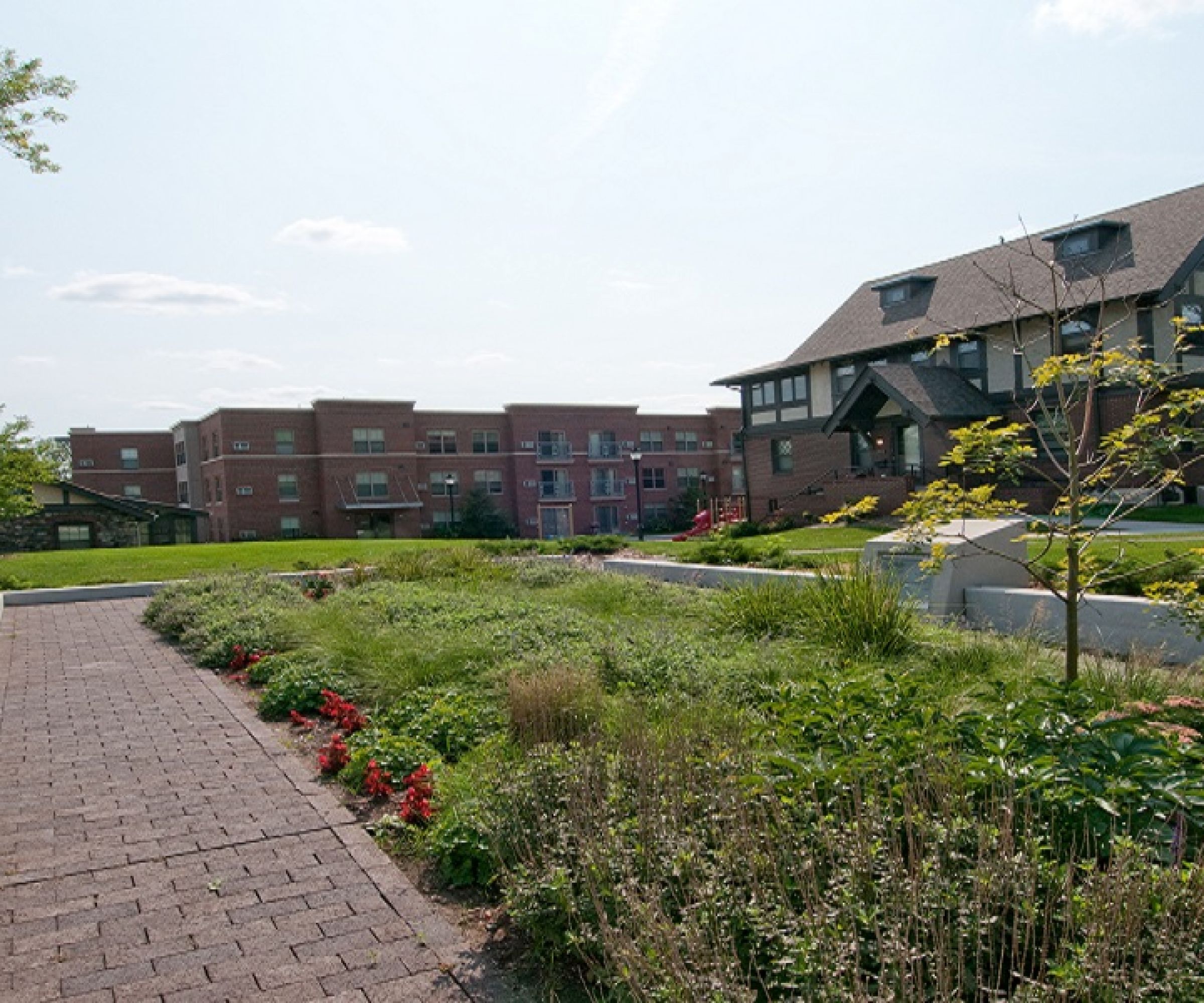 Aeon Affordable Homes and Community A walkway through a garden with benches leads to affordable apartment homes in charming brick and Tudor-style buildings under a partly cloudy sky.