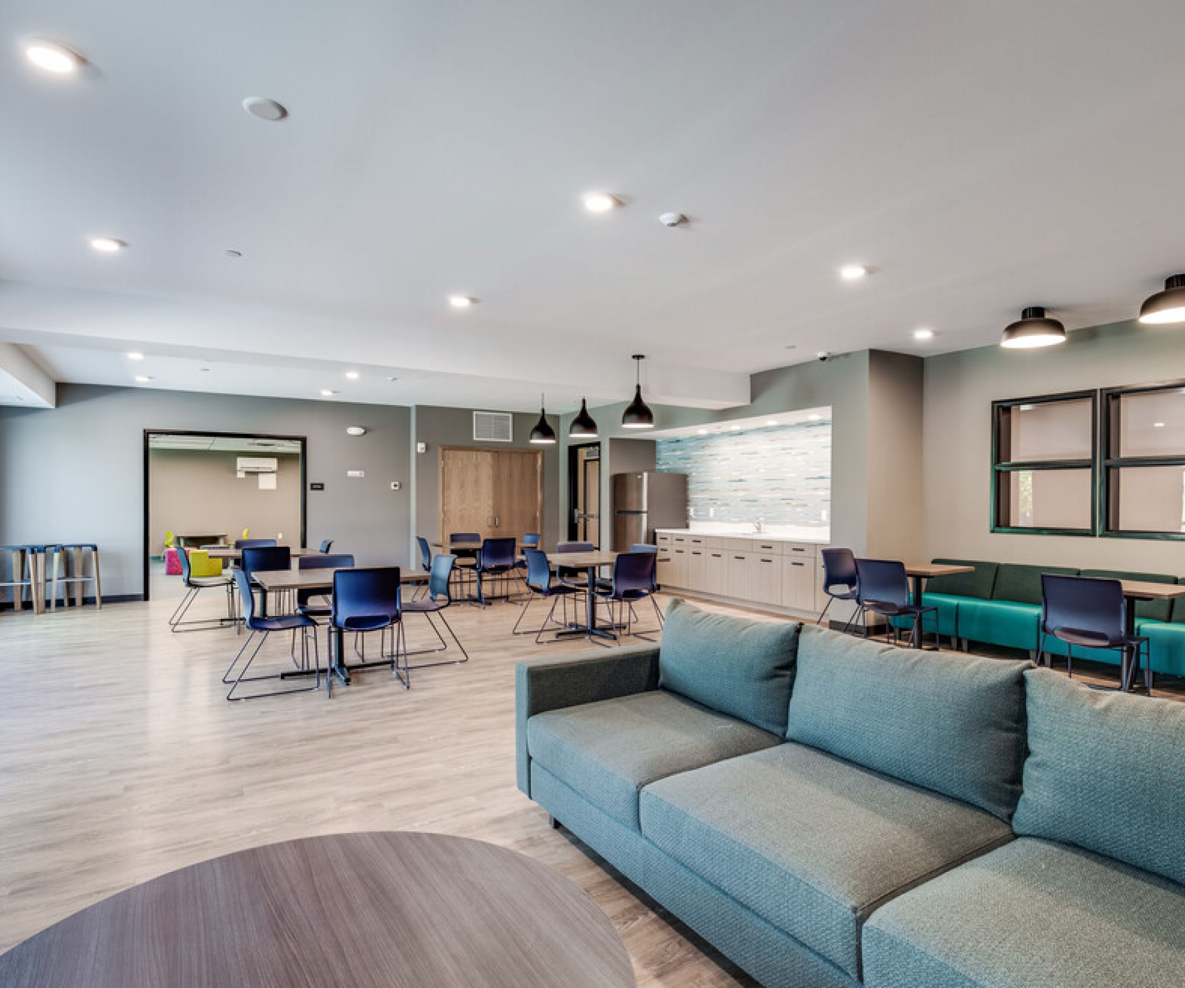 Aeon Affordable Homes and Community Modern lounge area with blue couches, tables, and chairs in affordable apartment homes, showcasing large windows for natural light.