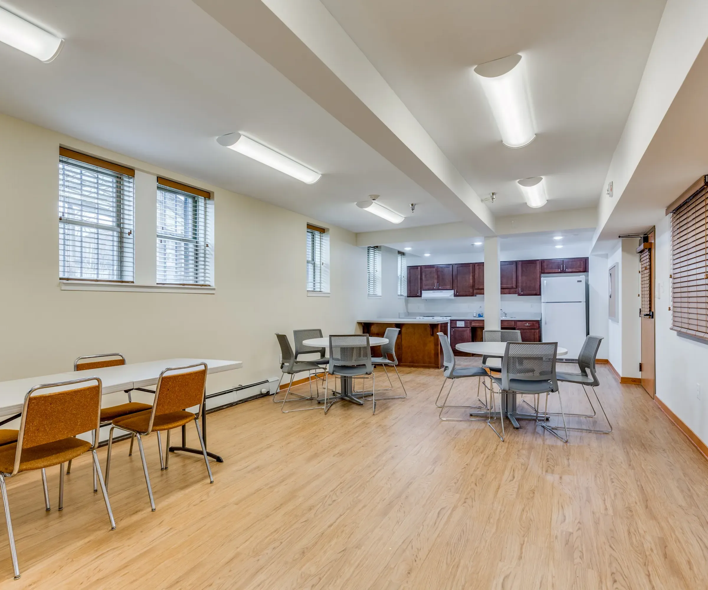 Aeon Affordable Homes and Community A bright, spacious room in affordable apartment homes, featuring tables, chairs, a wooden floor, and a kitchen area in the background.