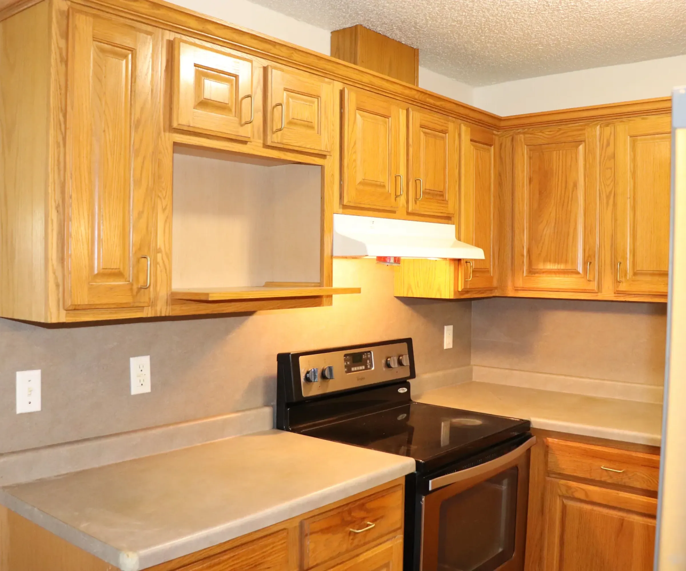 Aeon Affordable Homes and Community Affordable apartment home kitchen with wooden cabinets, beige countertops, black stove, and white range hood. Refrigerator partially visible.
