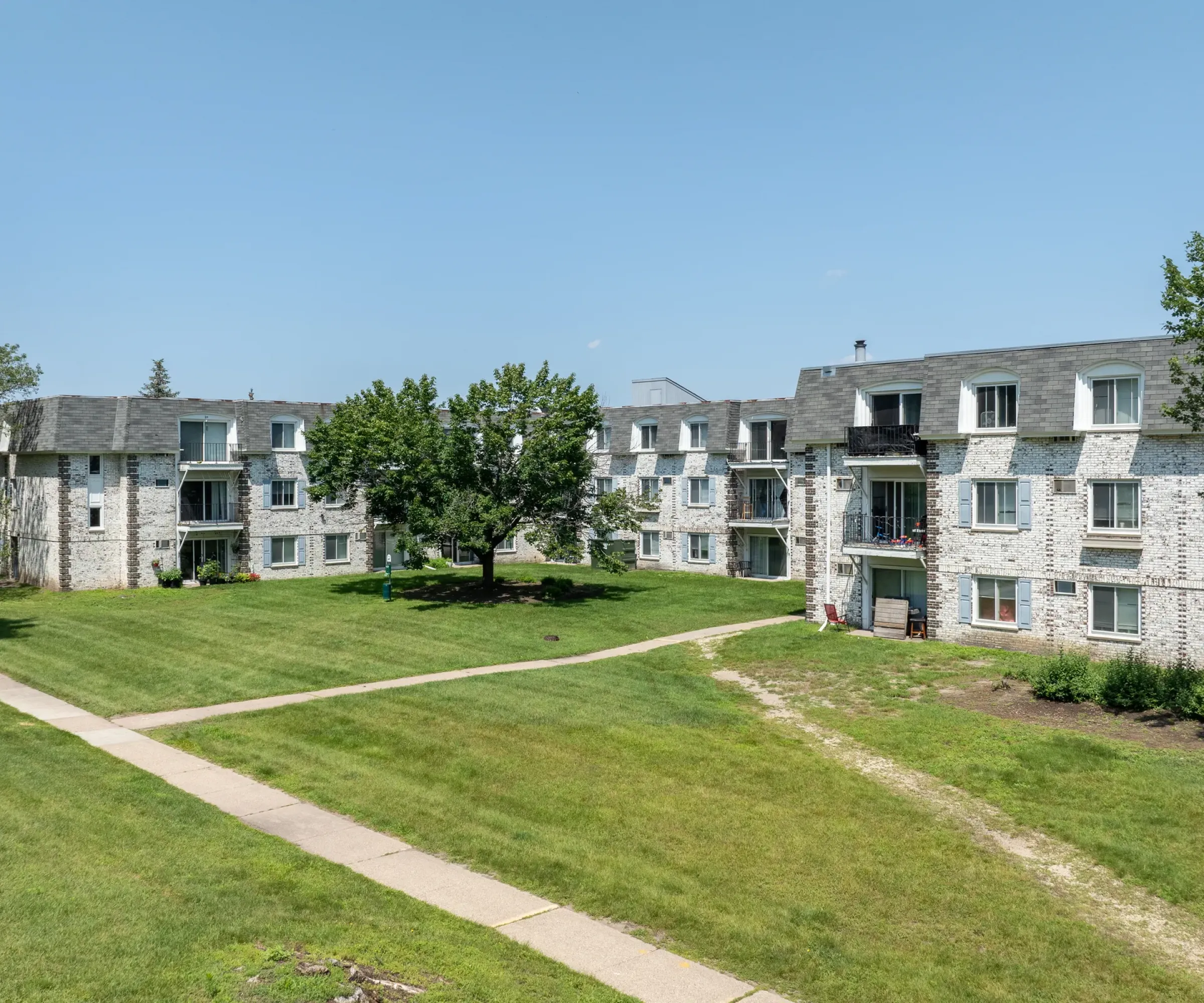 Aeon Affordable Homes A large, grassy courtyard in Blooming Meadows is surrounded by stone apartment buildings under a clear blue sky.