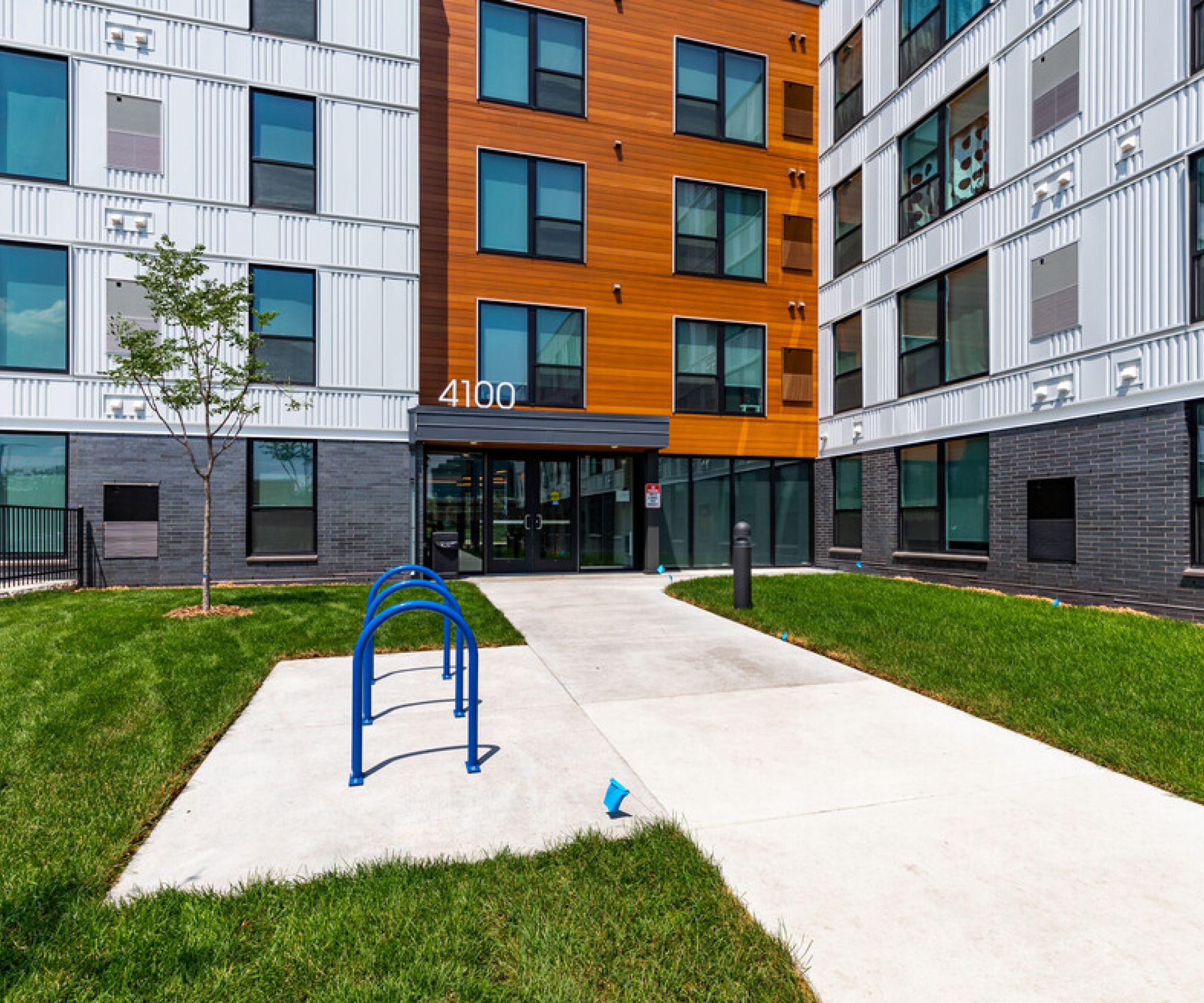 Aeon Affordable Homes and Community Affordable apartment homes featuring a modern entrance, with a blue bike rack, surrounded by green grass and a small tree.