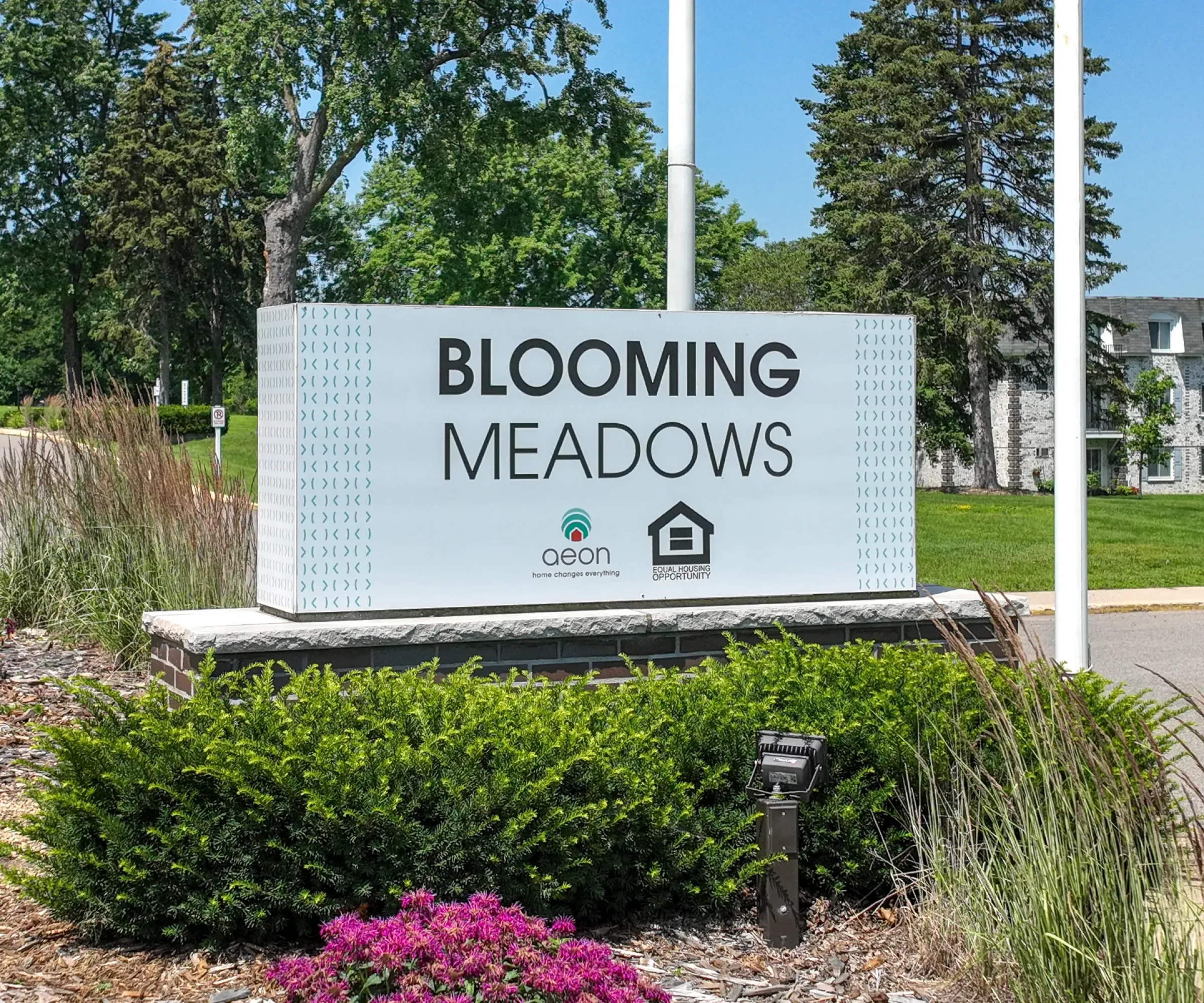 Aeon Affordable Homes A sign reading "Blooming Meadows South" stands amidst lush greenery, with charming buildings in the backdrop.