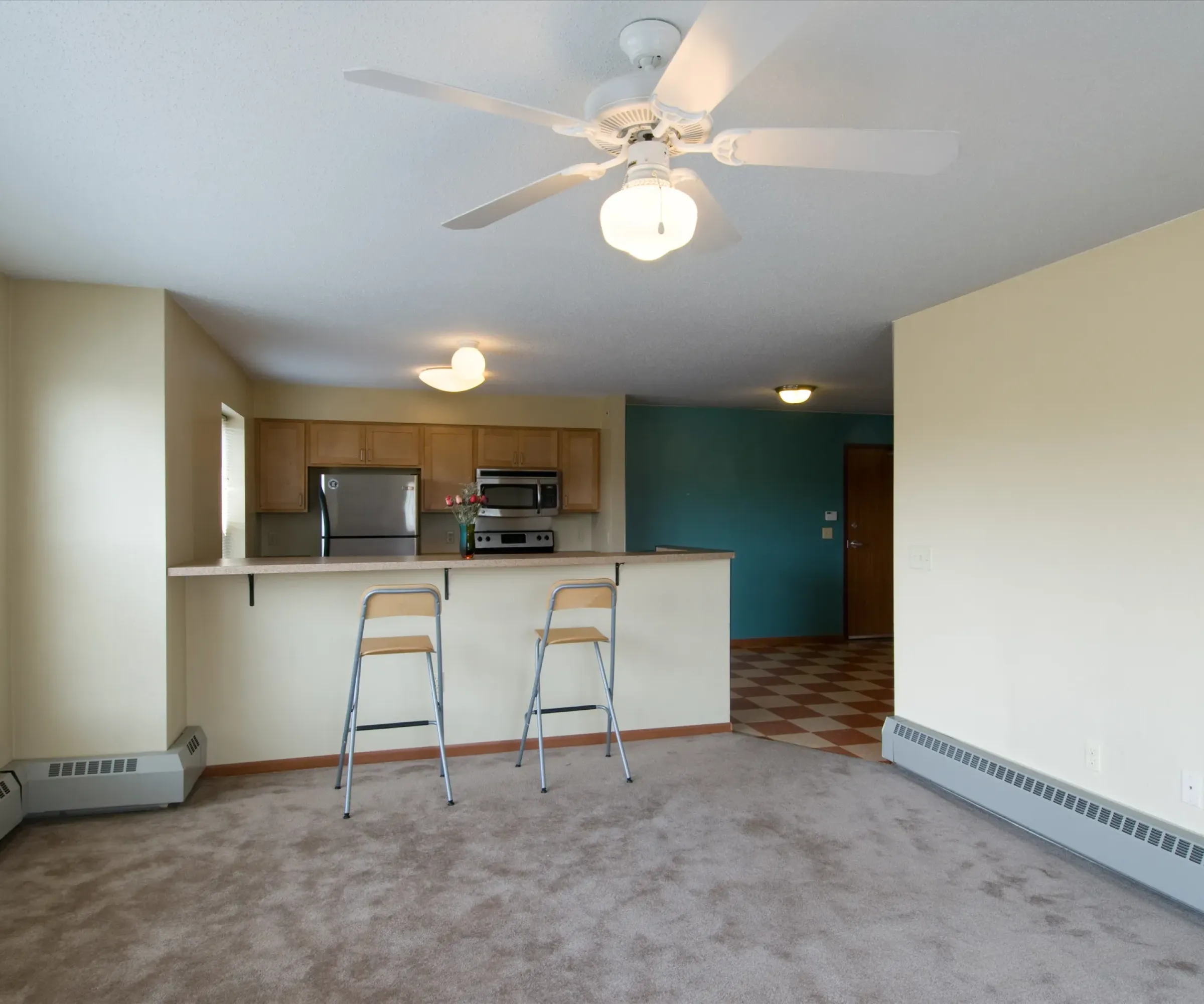 Aeon Affordable Homes and Community Bright, affordable apartment homes with an empty living room featuring beige carpet, bar stools, ceiling fan, and a view of the kitchen's two-tone walls.