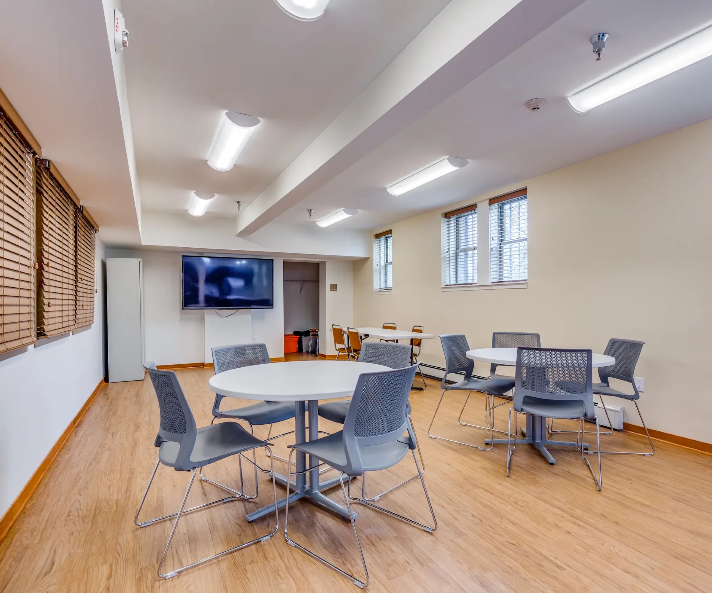 Aeon Affordable Homes and Community A meeting space with circular tables, gray chairs, a large TV on the wall, and wooden flooring, echoing the style of affordable apartment homes.