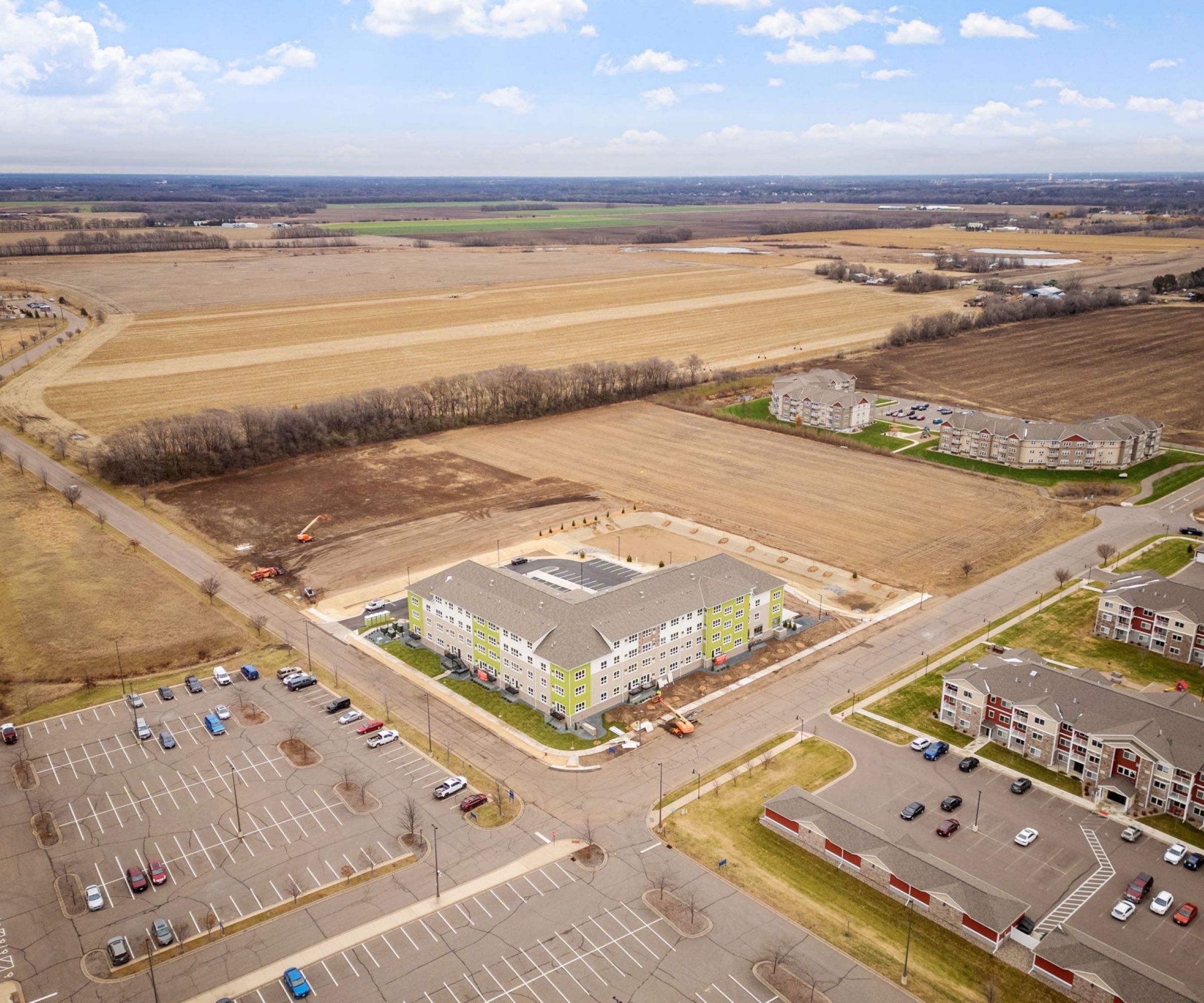 Aeon Affordable Homes and Community Aerial view of affordable apartment homes, parking lots, residential buildings, and vast open fields under a partly cloudy sky.