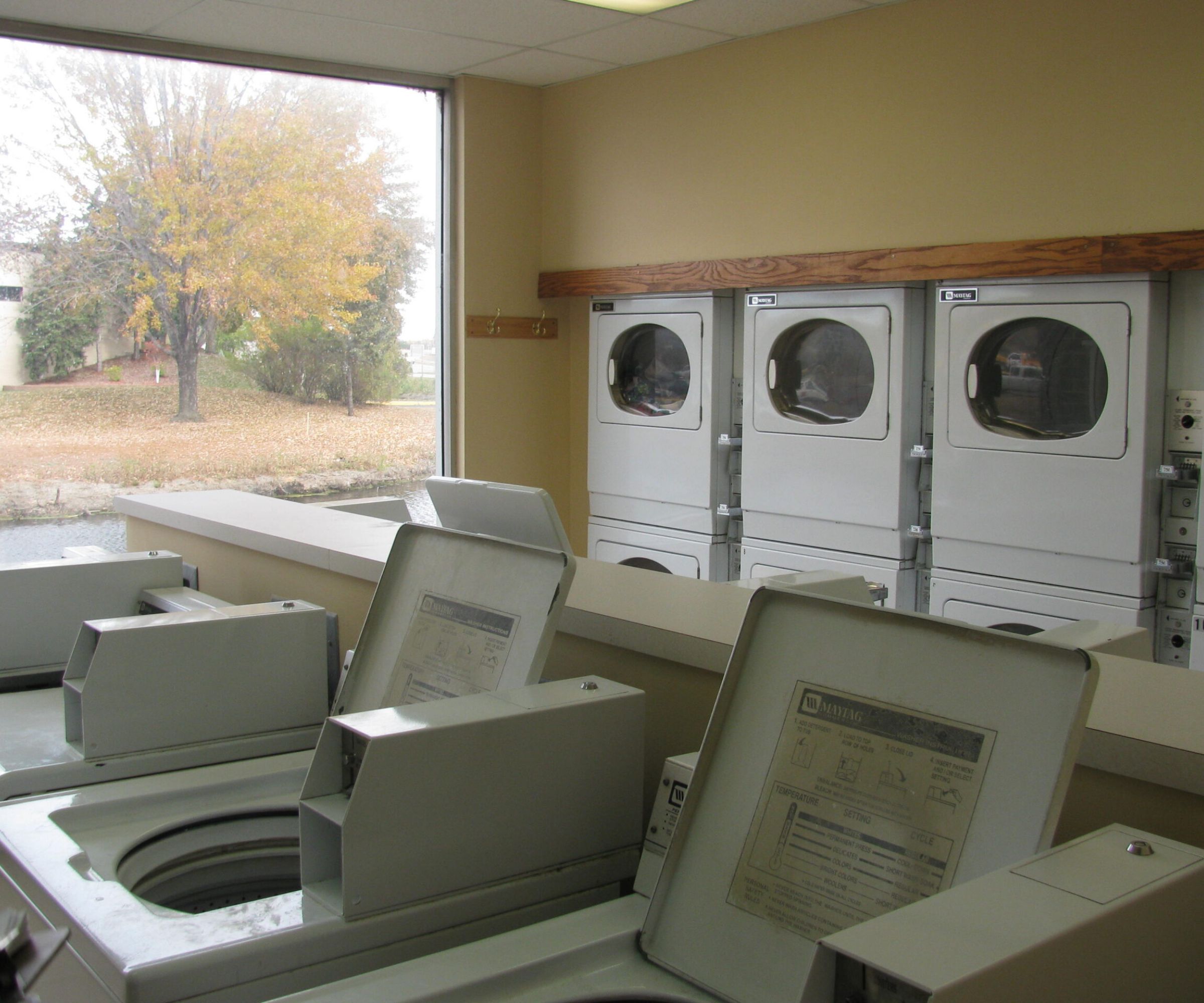 Aeon Affordable Homes and Community Interior of a laundromat with open washing machines and stacked dryers by a window, reminiscent of the view from affordable apartment homes.
