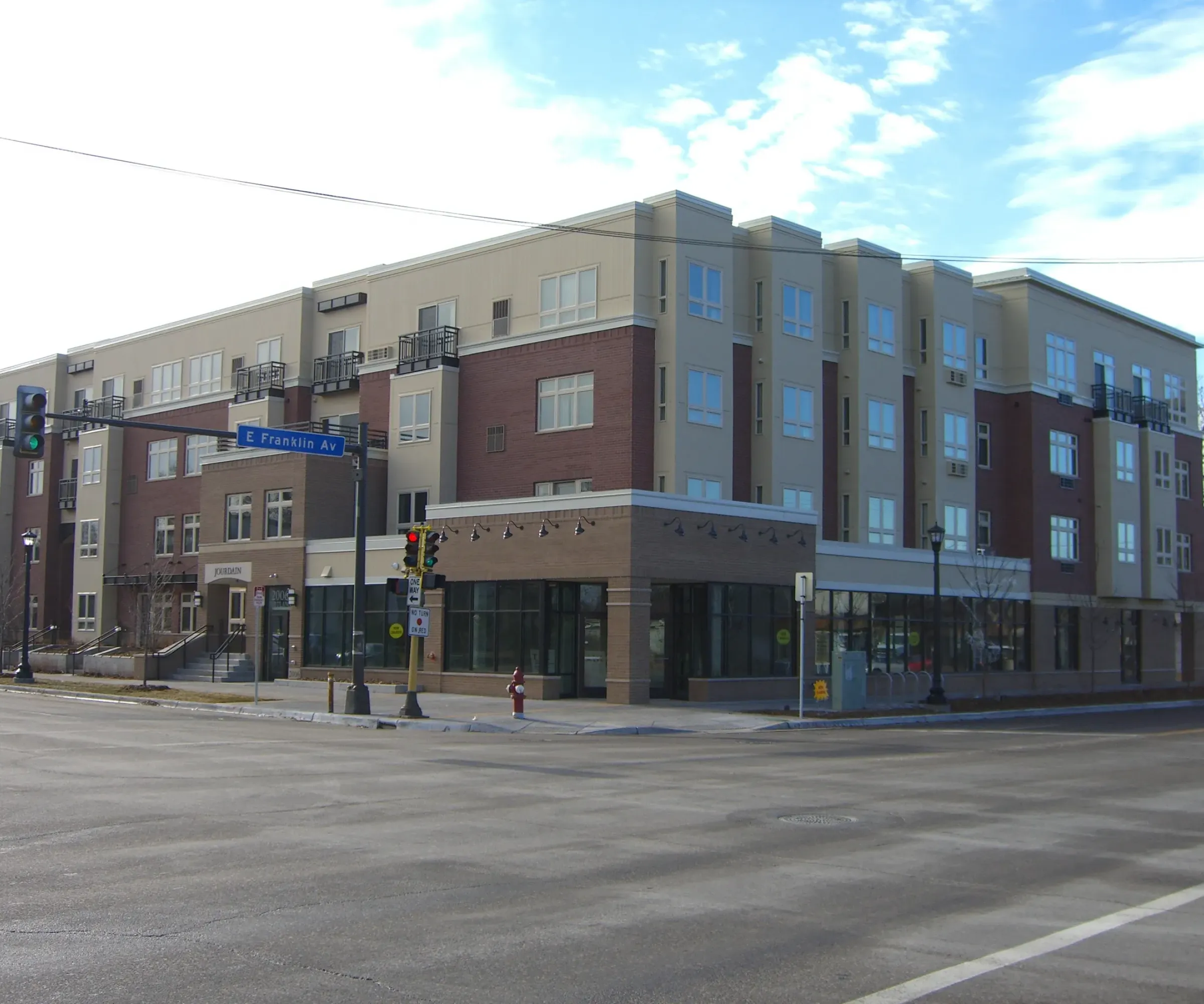 Aeon Affordable Homes and Community A four-story building offering affordable apartment homes stands at an intersection under a partly cloudy sky, with a streetlight and road signs visible.