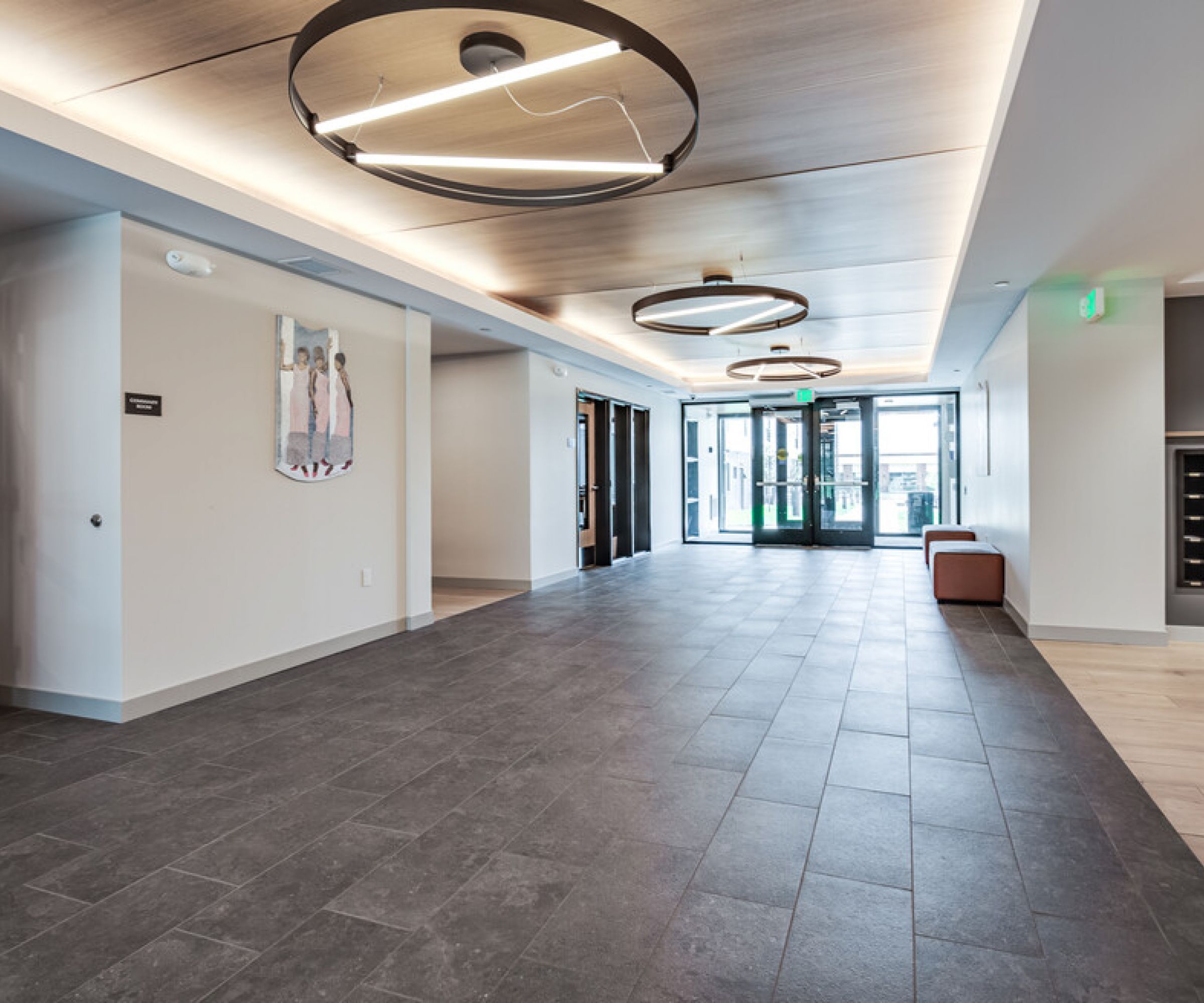Aeon Affordable Homes and Community Modern lobby with large circular lights, gray tile floor, wall art, and glass doors leading outside to affordable apartment homes.