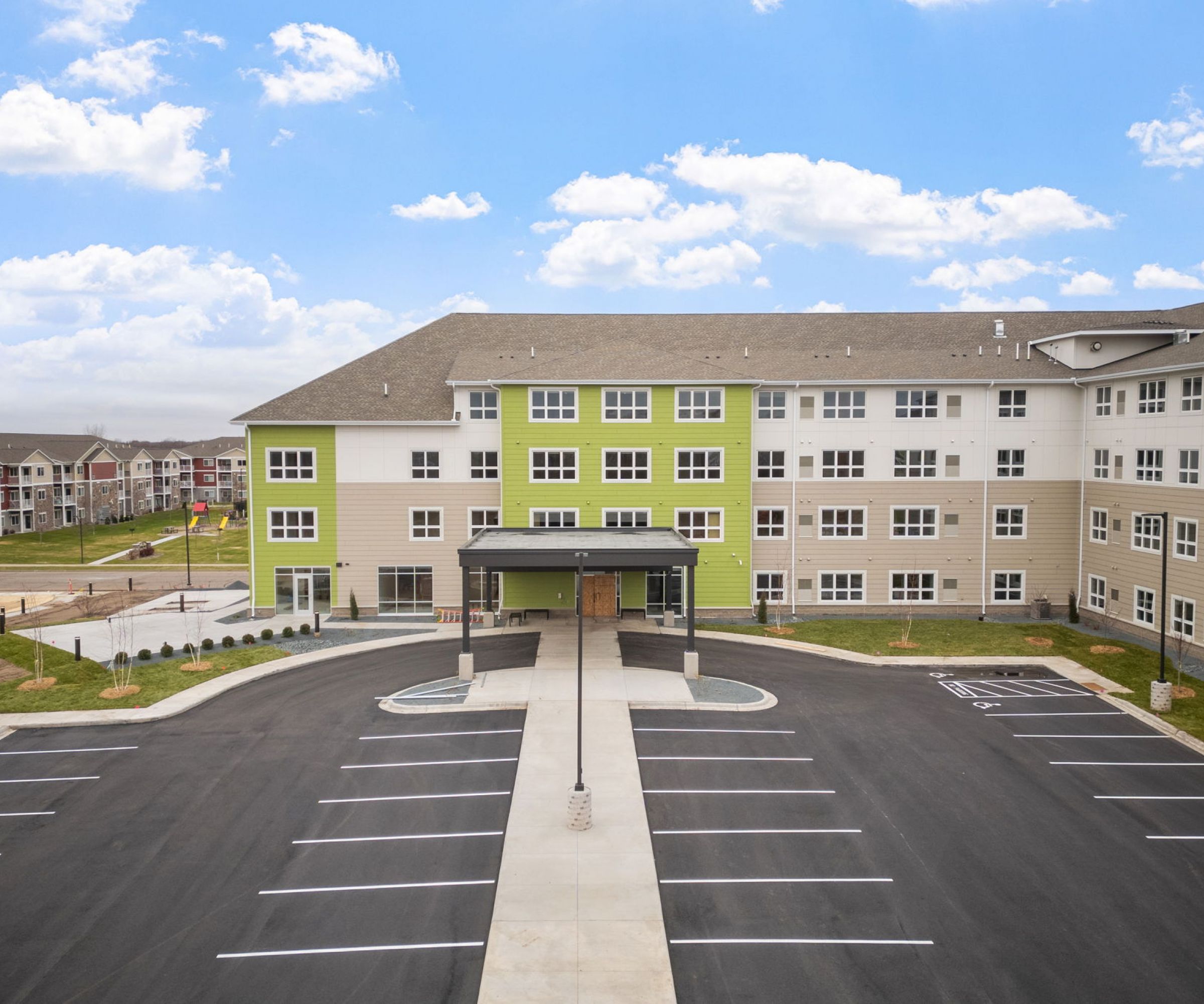 Aeon Affordable Homes and Community Large, four-story building with a green accent offering affordable apartment homes, surrounded by an empty parking lot and partly cloudy sky.