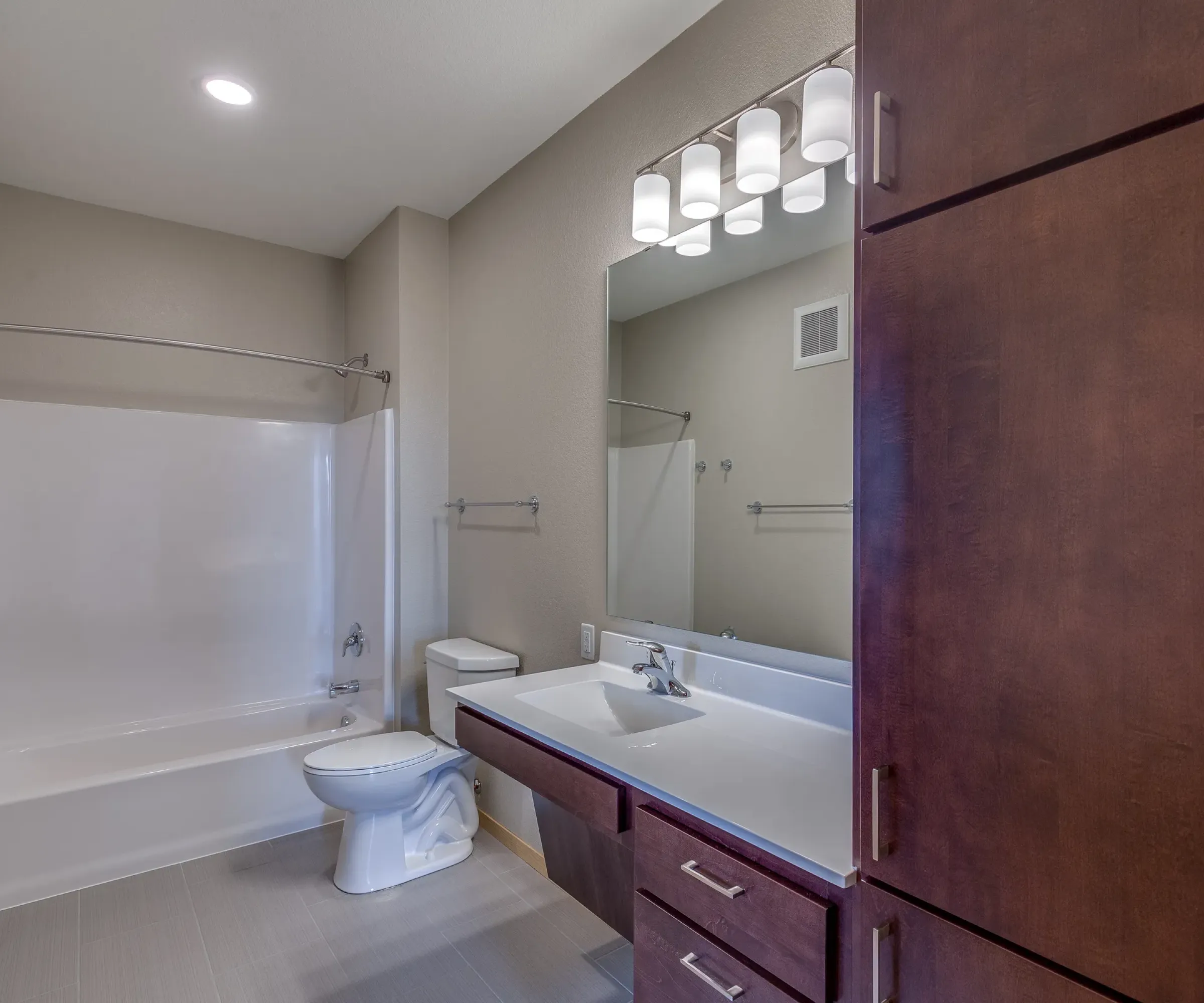 Aeon Affordable Homes and Community Affordable apartment homes featuring a modern bathroom with a shower-tub combo, white vanity, large mirror, and brown cabinets under soft lighting.