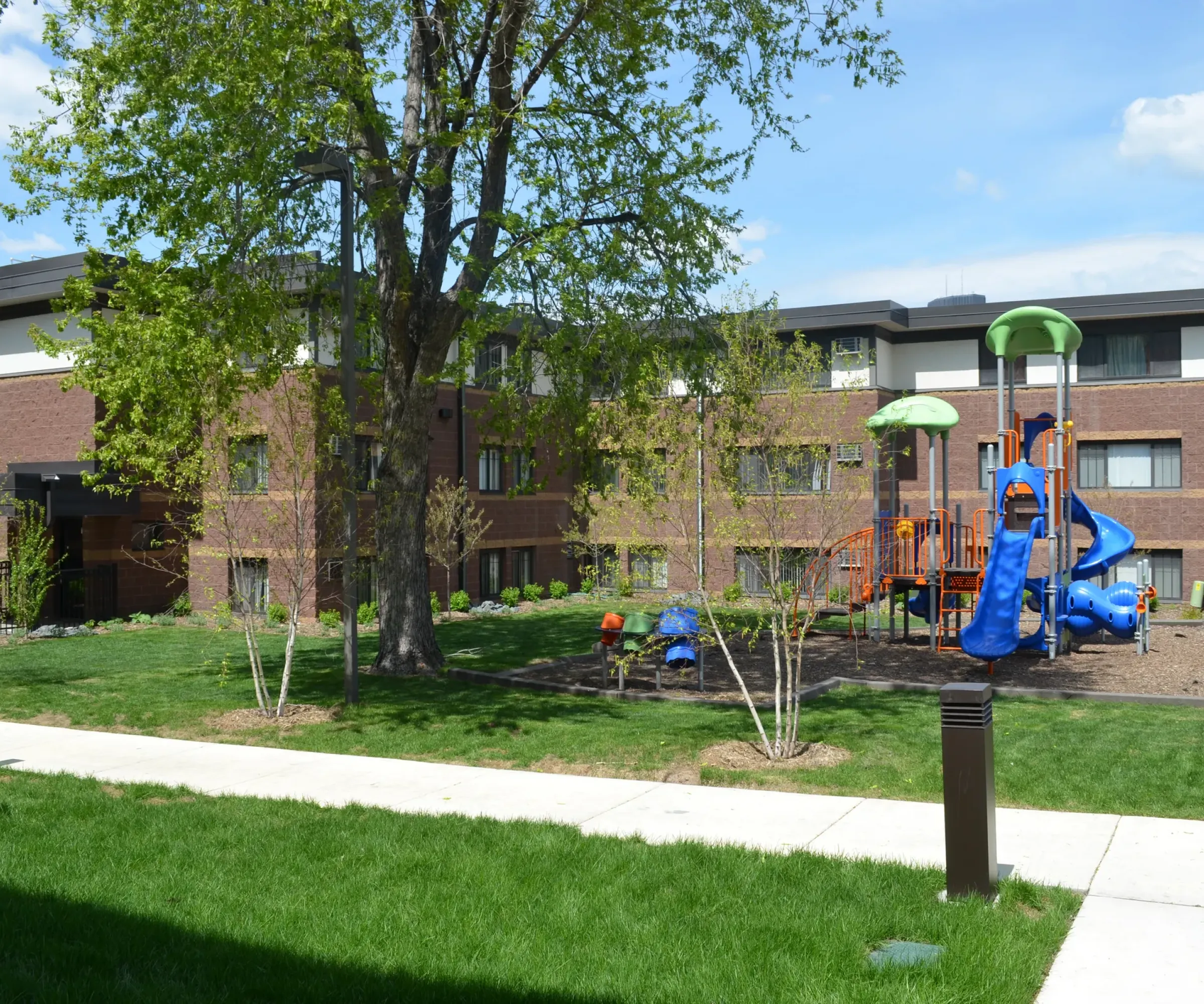 Aeon Affordable Homes and Community Next to affordable apartment homes, a playground features blue slides and climbing structures, surrounded by grass and trees.