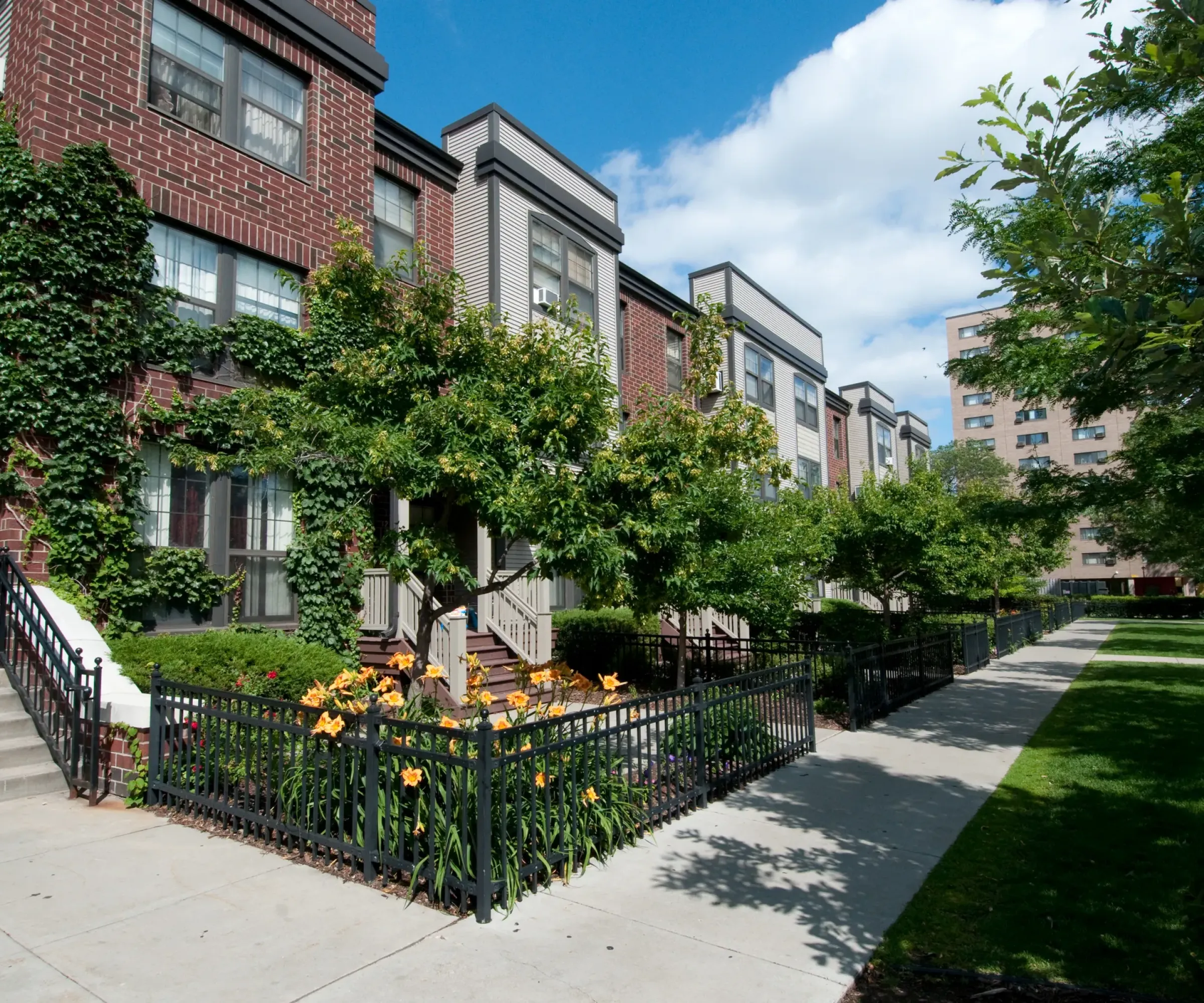 Aeon Affordable Homes and Community Brick row houses with lush green trees and orange flowers lining the sidewalk offer affordable apartment homes under a bright blue sky.