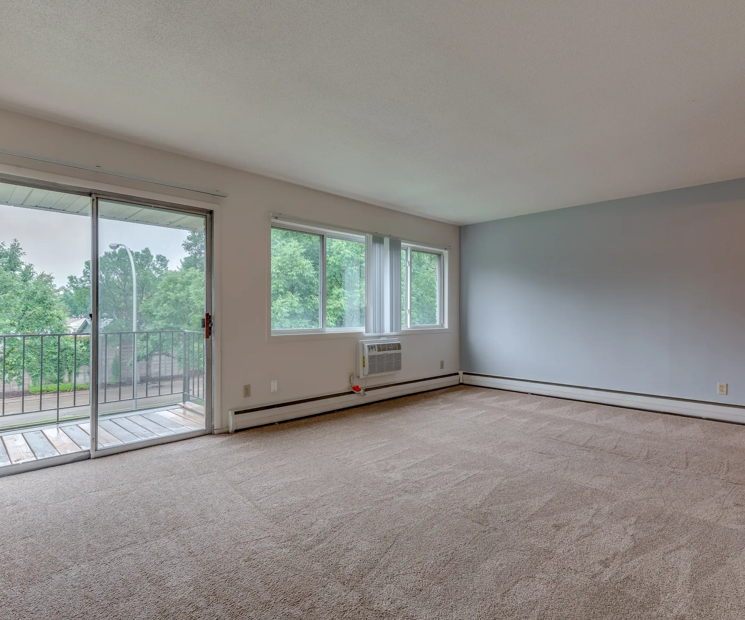 Aeon Affordable Homes and Community Empty carpeted room with large windows, sliding glass door to a balcony, and neutral walls in our affordable apartment homes.