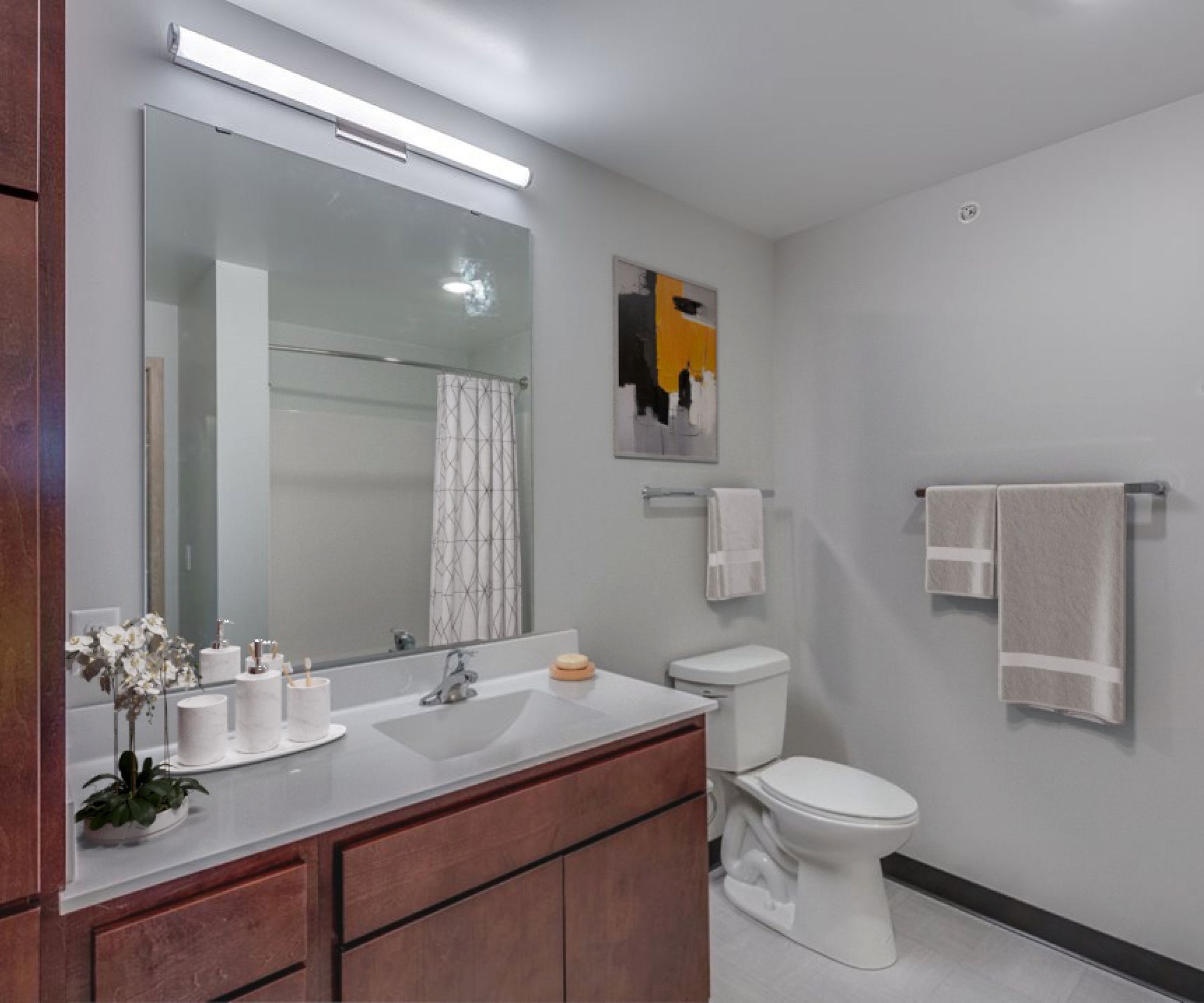 Aeon Affordable Homes and Community Modern bathroom in affordable apartment homes featuring a large mirror, wooden vanity, towels, and a decorative painting on a light gray wall.