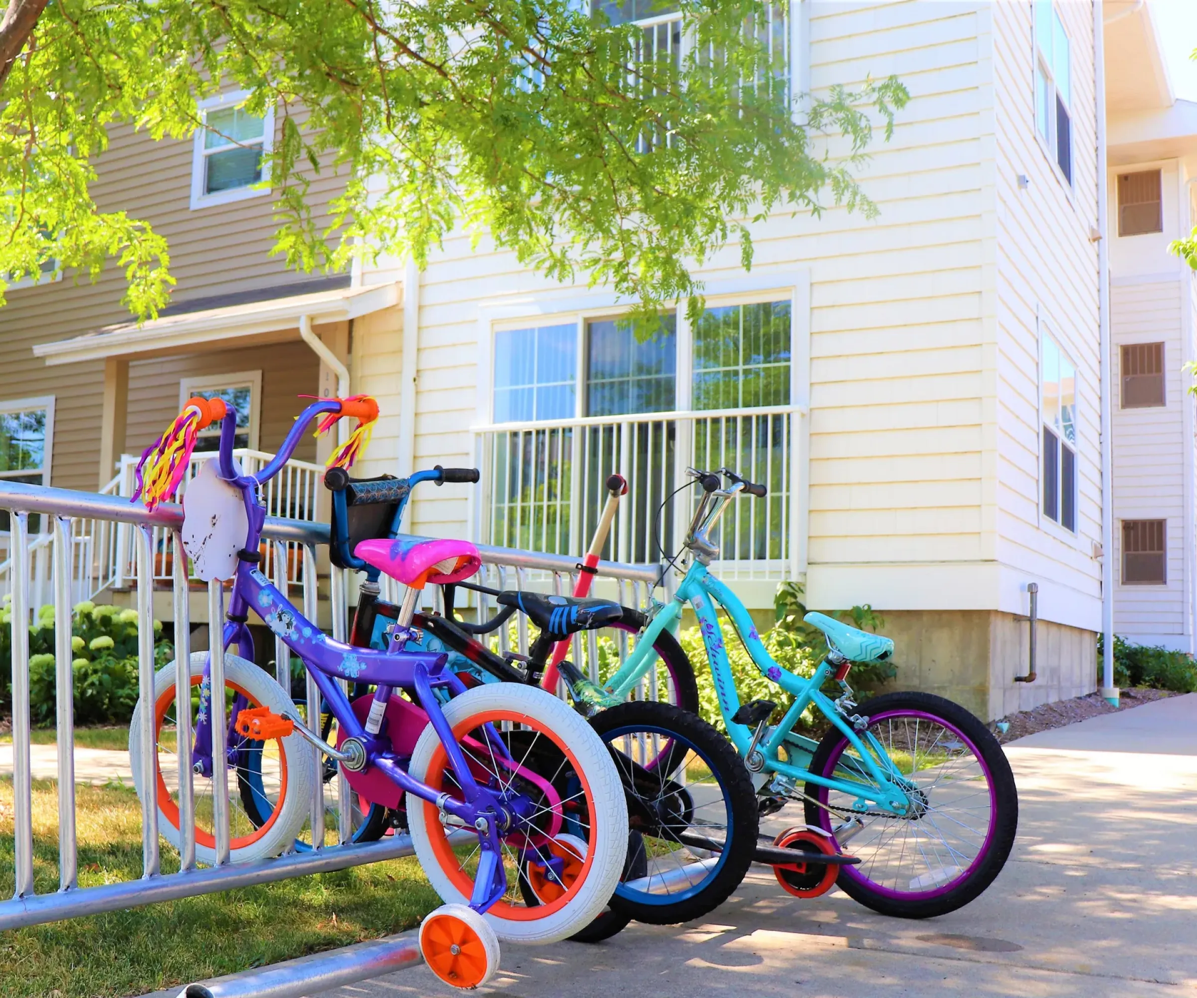 Aeon Affordable Homes and Community Four colorful children's bicycles parked near an affordable apartment home, beside a tree and a light-colored building.