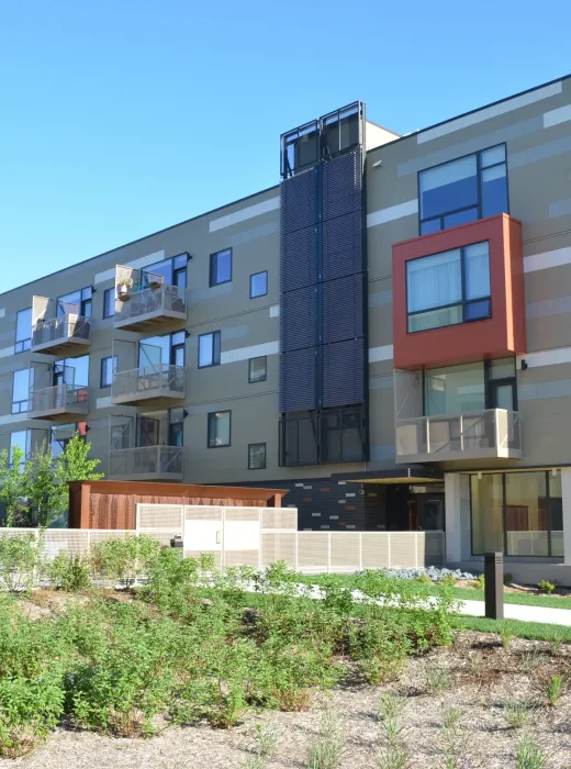 Aeon Affordable Homes and Community Modern, affordable apartment homes with balconies in a four-story building, surrounded by landscaped greenery under a clear blue sky.