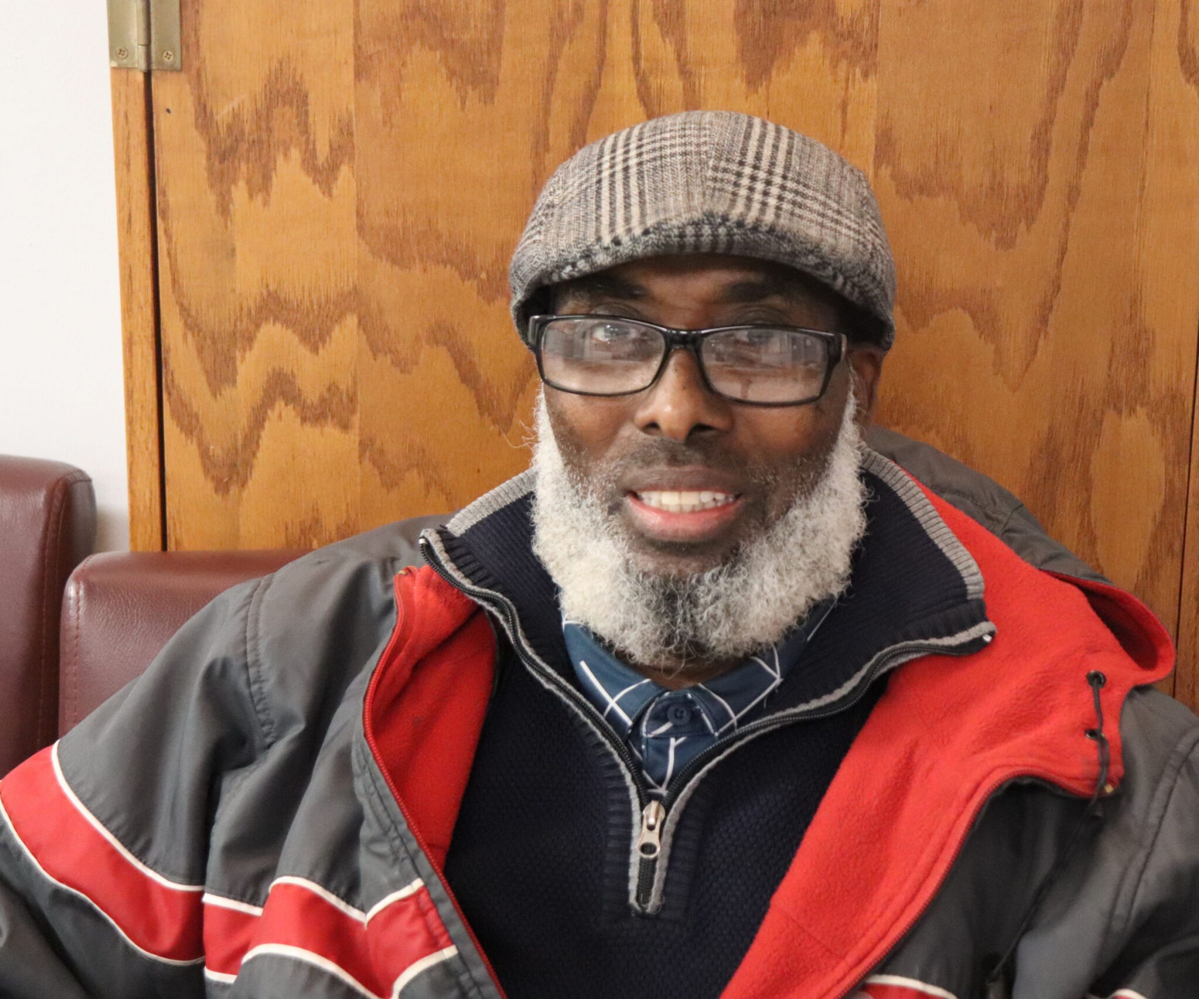 Aeon Affordable Homes A smiling man with a gray beard, glasses, and a flat cap sits on a maroon chair in a jacket, embodying Jama's Story.