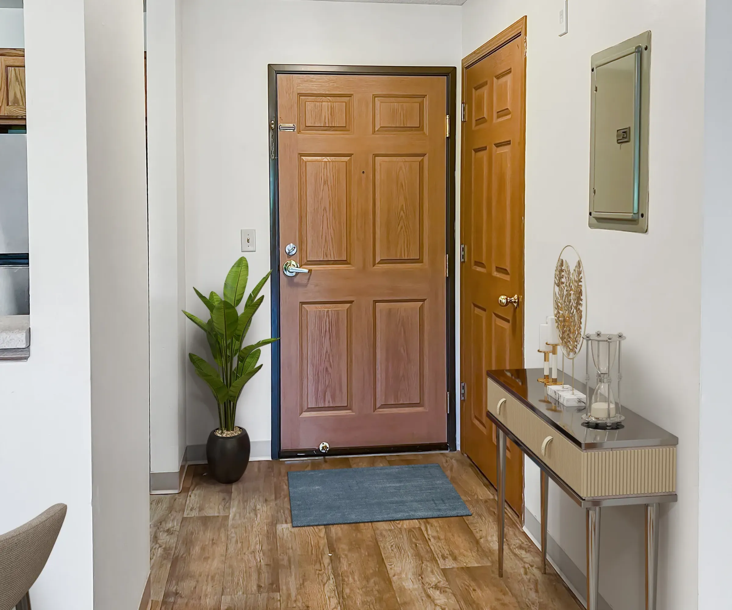 Aeon Affordable Homes and Community Entryway of affordable apartment homes with a wooden door, potted plant, small table, and blue rug on wood flooring. Bright lighting overhead.