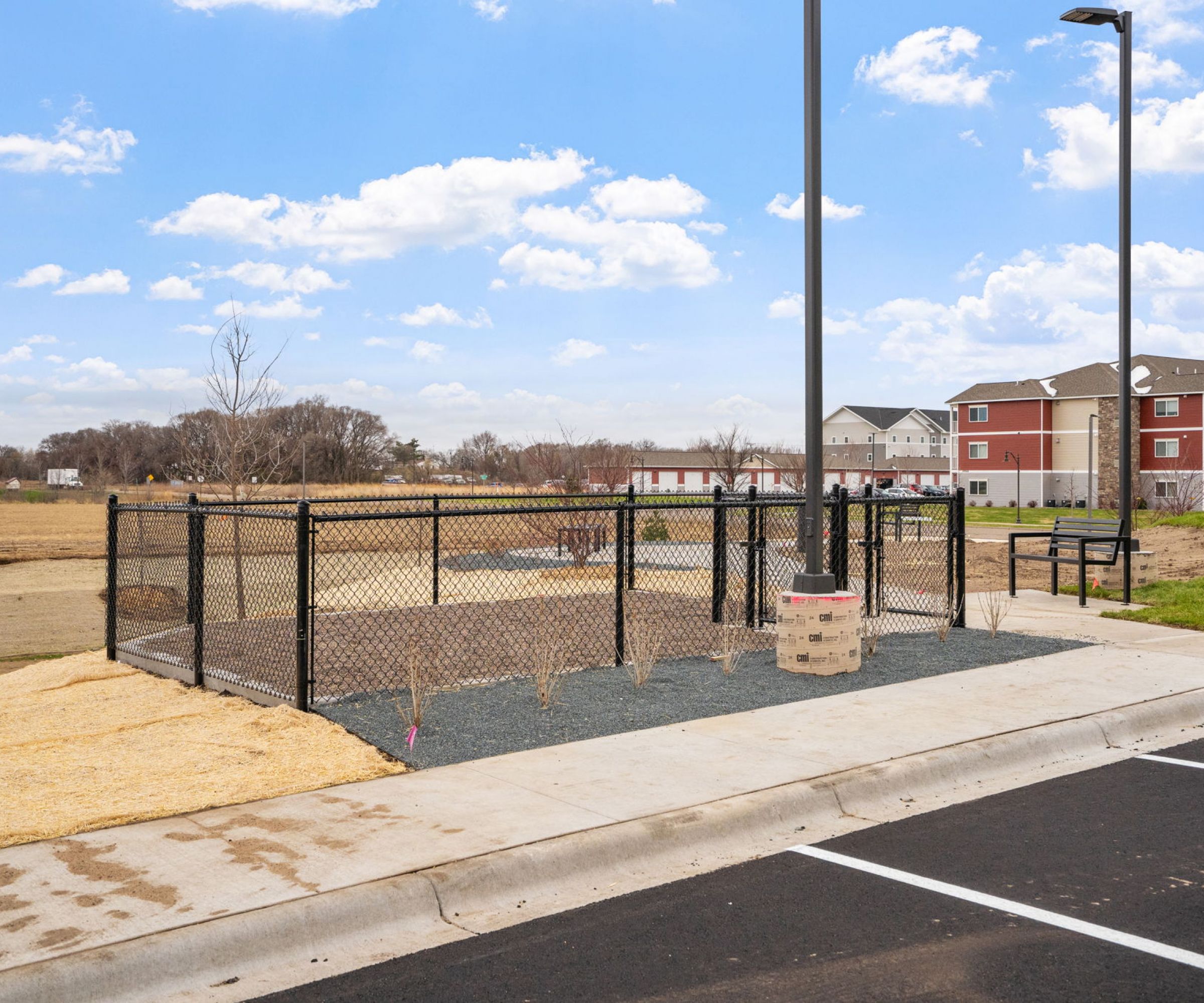 Aeon Affordable Homes and Community Small fenced dog park near parking lot and affordable apartment homes under a partly cloudy sky.