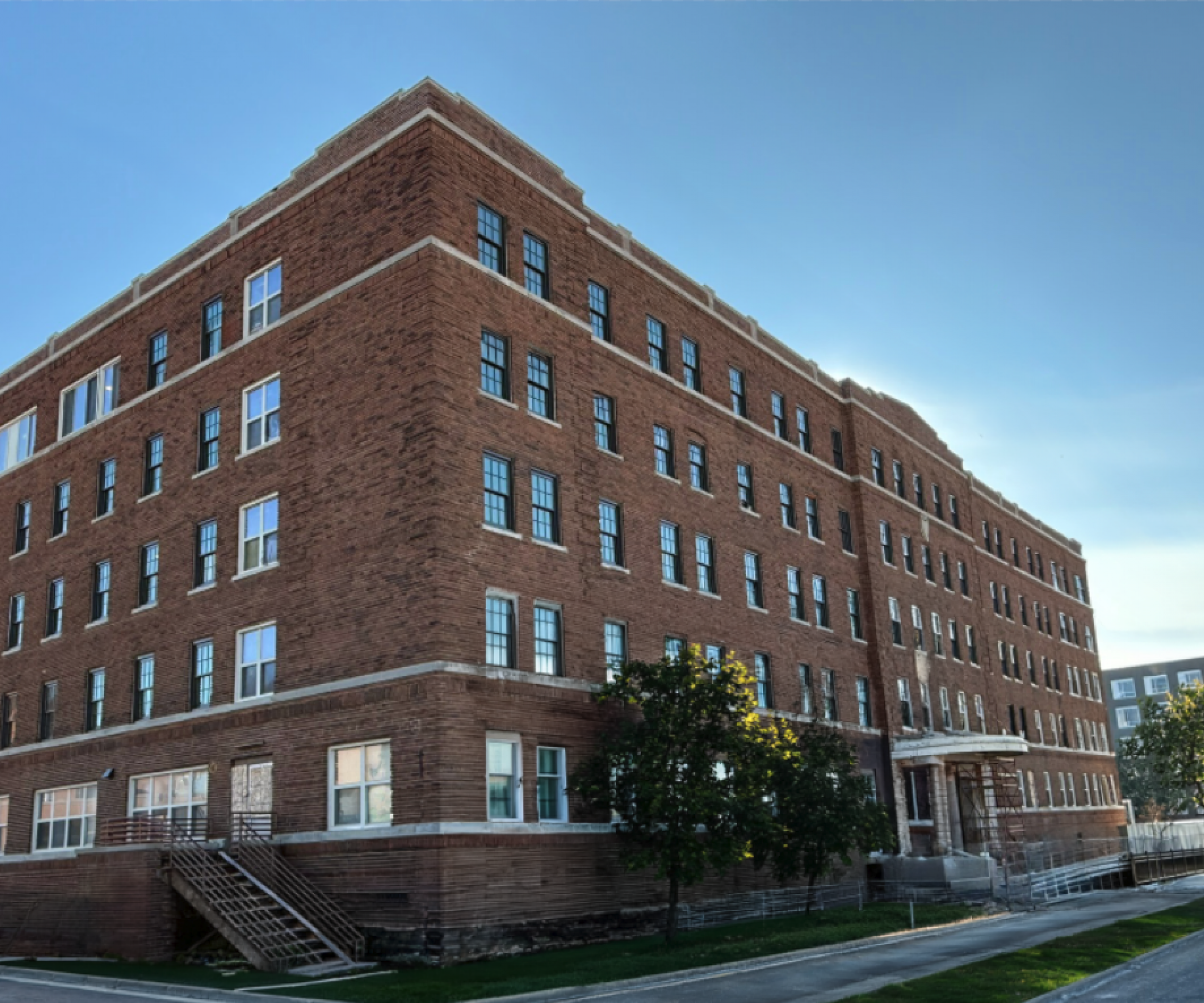Aeon Affordable Homes A large, five-story red brick building on a sunny day with trees and sidewalks along its edge.